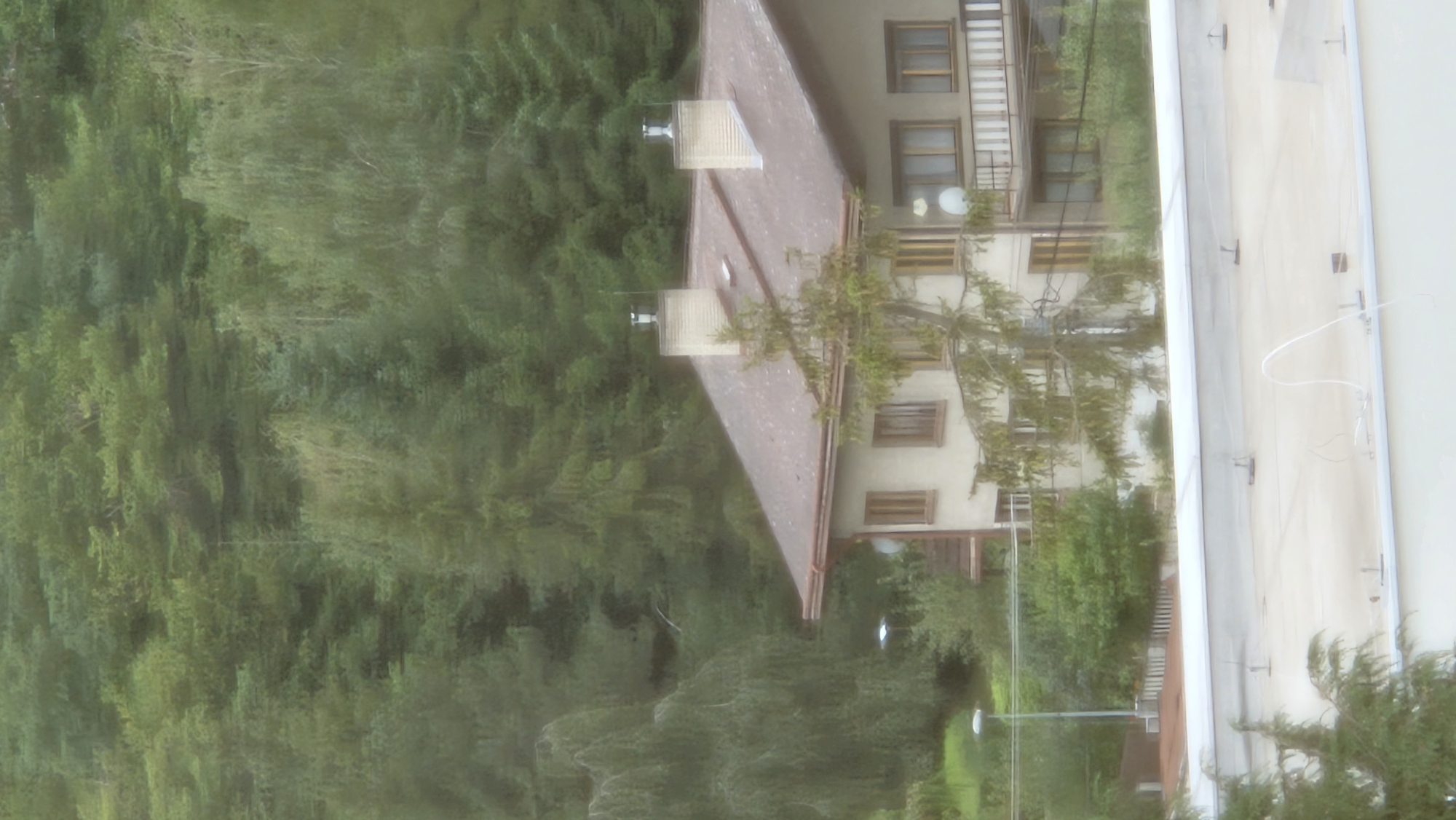 The image shows a residential building with a reddish-brown roof, flanked by trees and foliage. The building has multiple windows and two chimneys on the roof, each topped with a metallic vent. A tall streetlamp is visible to the left of the building. The building appears to be situated on a slightly elevated slope, with a dense forest forming the backdrop. The foreground shows a portion of another building's roof, with what appears to be wiring and some fixtures attached to it. The overall scene suggests a semi-urban or suburban environment with significant natural elements. The specific location in Prešov, Slovakia, is not discernible from the visual information alone.