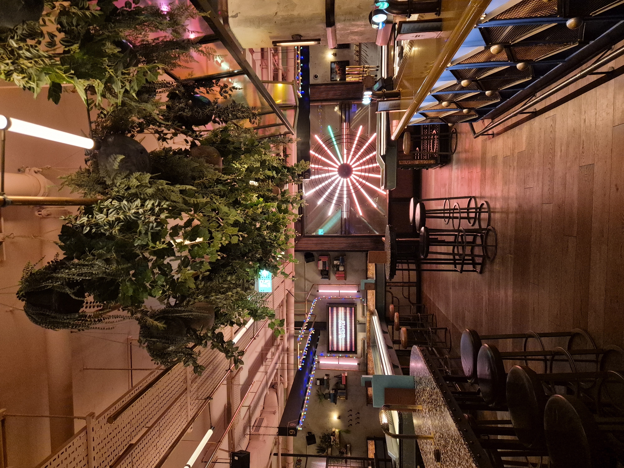 The image shows an interior view of a building, likely a bar or restaurant, possibly in London, United Kingdom. The perspective is an aerial view looking down. The space is characterized by a mix of industrial and natural elements. On the left, a lush arrangement of greenery hangs down, intertwined with exposed piping and lighting fixtures. Towards the center, a circular neon light fixture with radial lines dominates the ceiling. Below, tables and chairs are arranged along the length of the space, with a bar counter visible on one side. The overall ambiance is dimly lit with warm tones, suggesting a relaxed and potentially upscale setting.