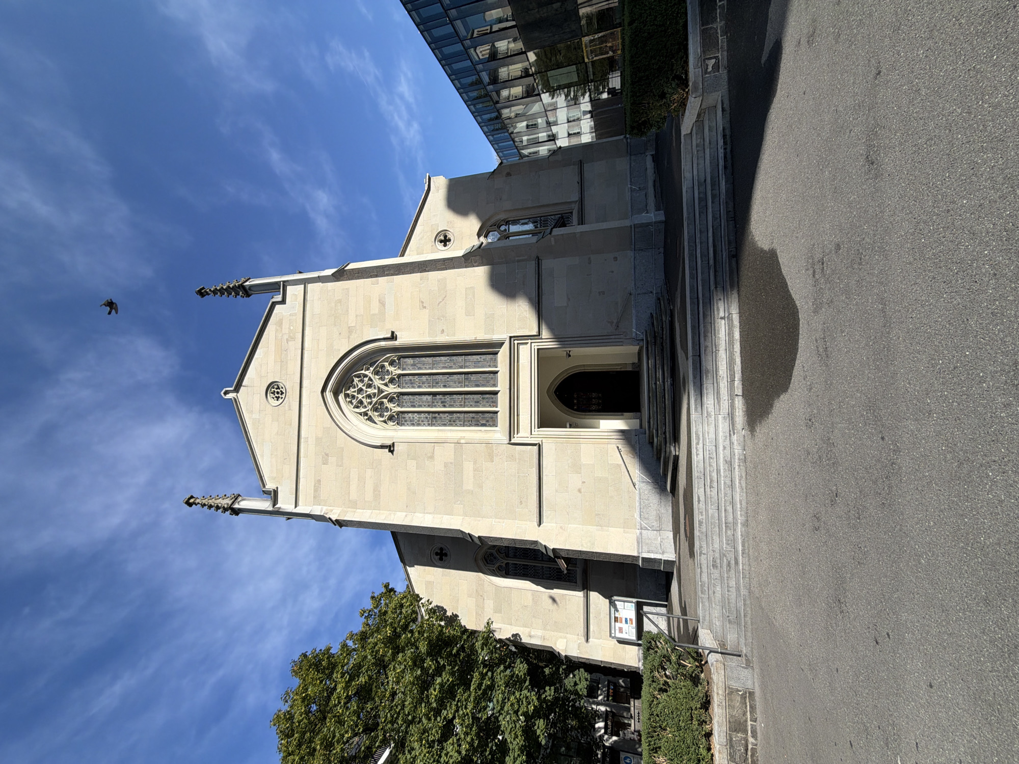 A low-angle, wide shot captures the front facade of a light-colored stone church under a bright blue sky with scattered wispy clouds. A single bird is in flight in the upper right quadrant of the sky. The church features a large arched entrance with a dark double door, topped by a tall, ornate arched window with intricate stained glass. Two spired towers flank the central pediment, and smaller arched windows are visible on the sides. A shadow of the church's left side falls across its facade. To the left of the church, a large green tree with dense foliage casts a shadow, and a modern glass-fronted building stands to the right. A set of wide stone steps leads up to the church entrance, flanked by manicured hedges. The ground in front of the steps is paved with asphalt, with a large dark shadow stretching across it. A signpost is visible to the left of the steps.