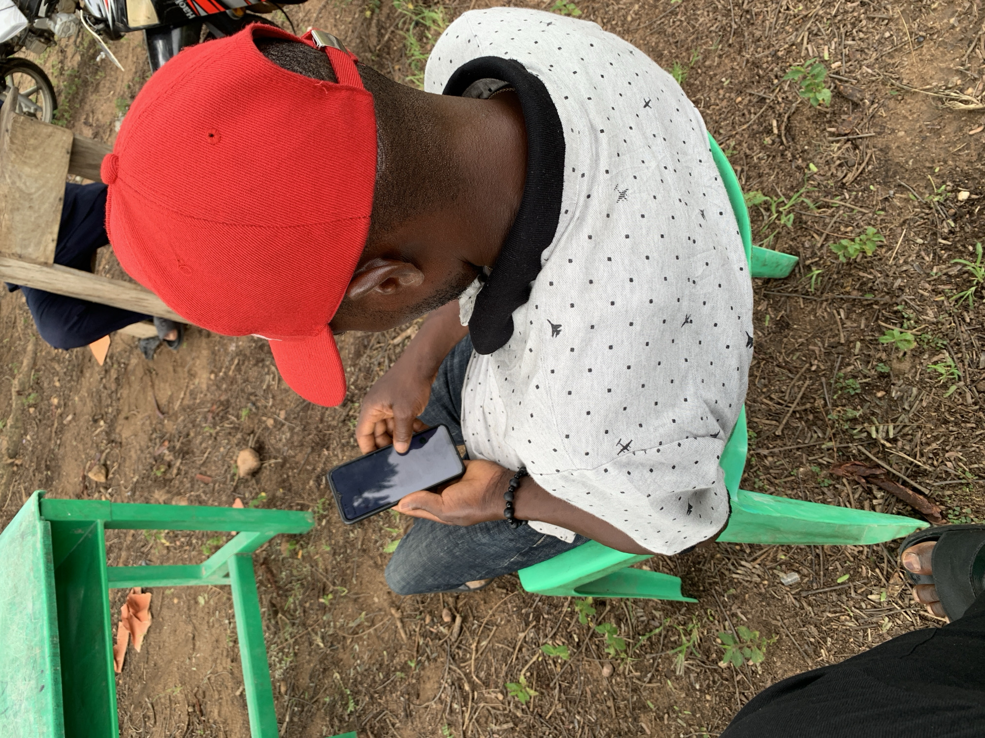 A high-angle, outdoor medium shot shows a dark-skinned man wearing a red baseball cap and a light gray shirt with a black speckled pattern sitting on a green plastic chair. He is holding a dark-colored smartphone in his right hand and appears to be looking at the screen. He is also wearing a black beaded bracelet on his left wrist. In the background, a green metal table with a shelf is visible, along with dry leaves and twigs on the ground. To the right of the man, part of a motorcycle is visible, suggesting an urban or semi-urban setting, possibly in Tabu, Nigeria, given the context provided. Another person wearing dark pants and sandals is seated behind a wooden structure, further to the left. The ground is composed of dirt and scattered debris.