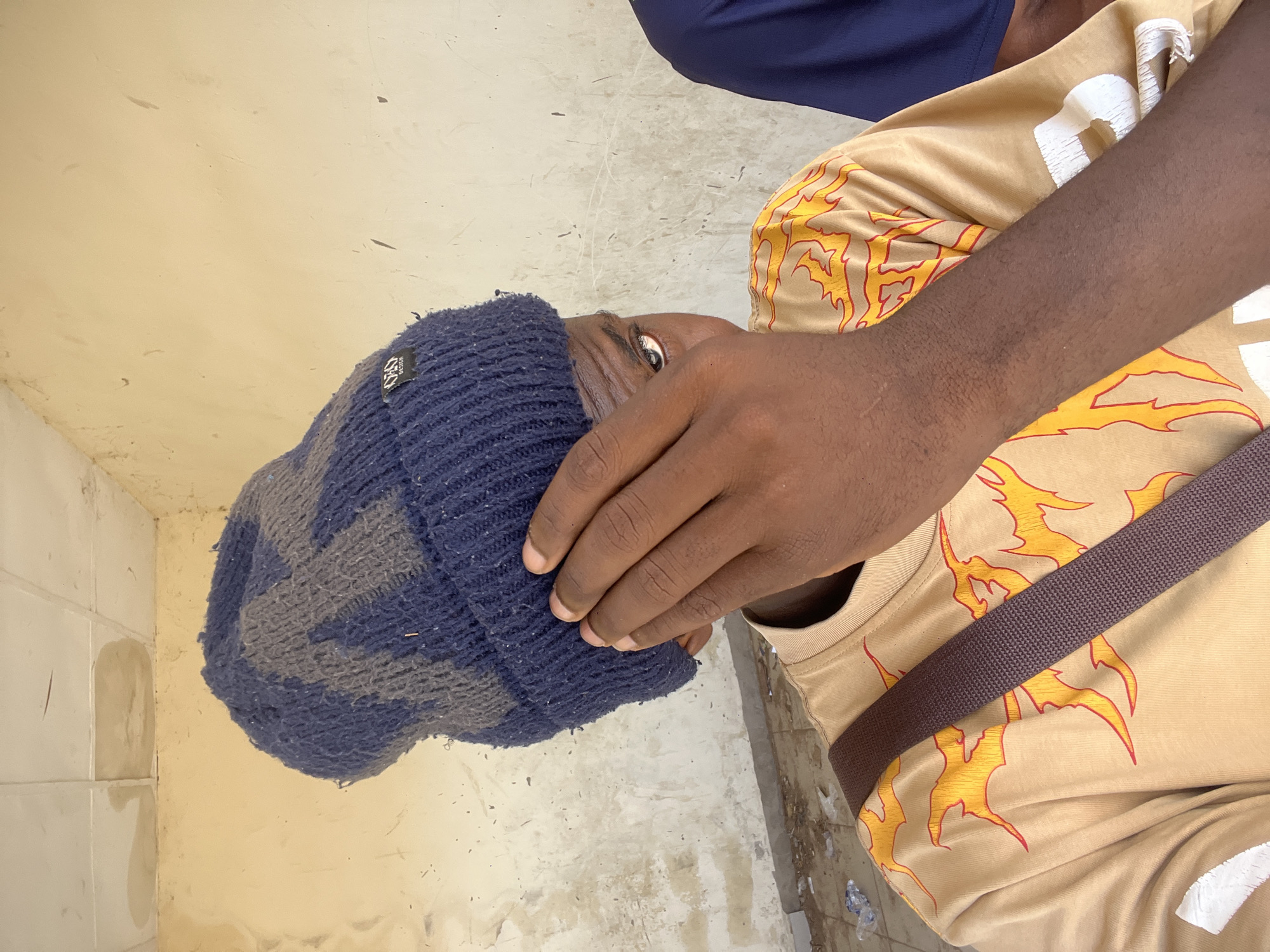 The image depicts a dark-skinned individual from the upper torso, observed within an indoor or covered area in Sabon Layi Jessu, Nigeria. The subject wears a dark blue and grey patterned knitted beanie, featuring a black rectangular tag inscribed "ORO DESIGN" on its left side. A light tan t-shirt adorned with stylized orange and yellow flame graphics covers the subject's body. A dark brown, textured strap, likely from a bag, diagonally crosses the chest from the upper left to the lower right. The subject's right hand is positioned over the lower portion of the face, obscuring the nose and mouth. The visible right eye, with a notable light reflection, looks directly towards the viewer. A segment of another individual, clad in a plain dark blue garment, is visible on the far right. The background features a weathered, light yellow wall exhibiting numerous dark smudges and vertical streaks. The ceiling consists of white acoustic tiles, several of which are severely discolored by extensive water damage and appear to be sagging. The floor in the lower left foreground is gritty and strewn with plastic debris and dirt.