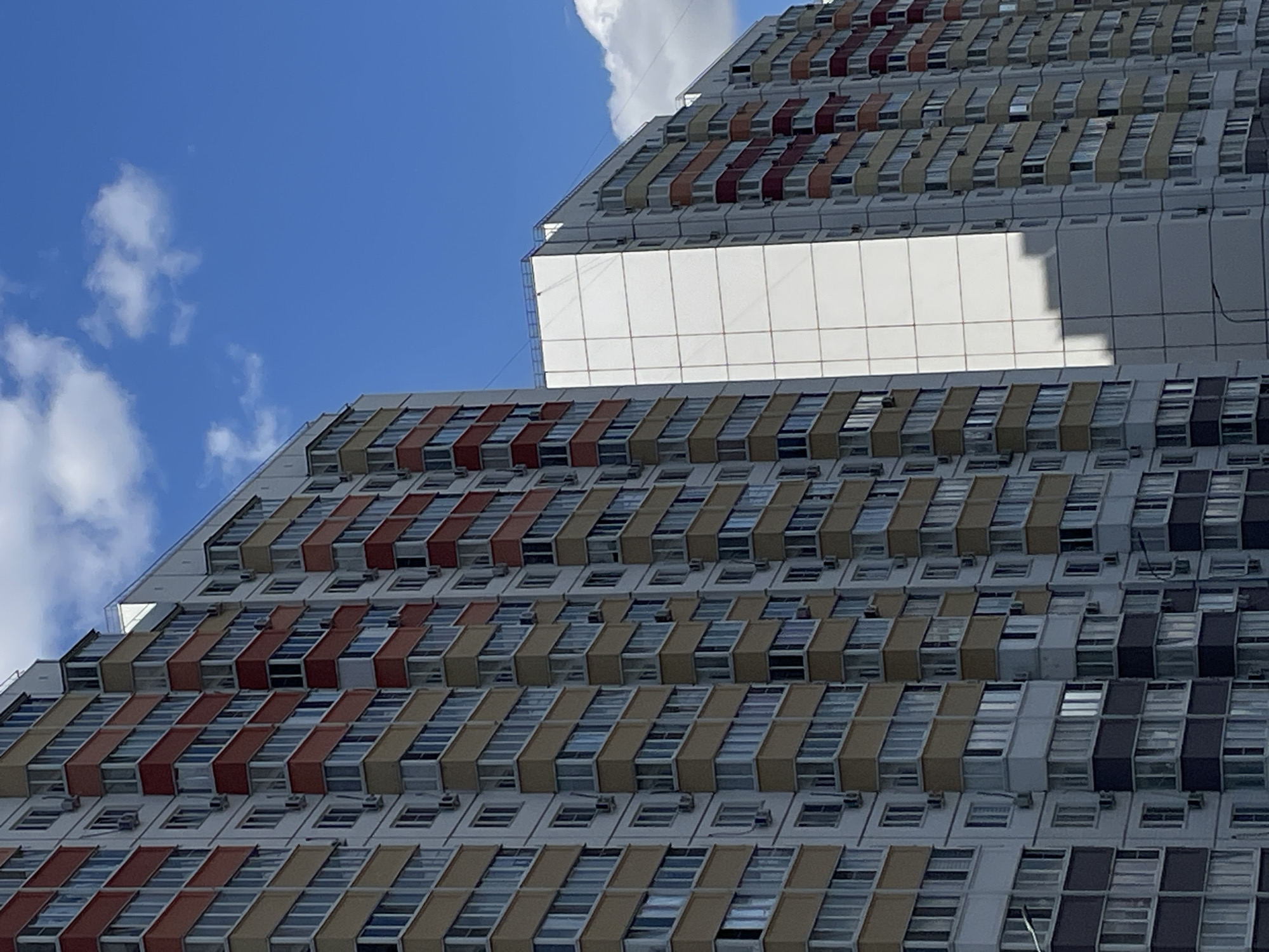 The image shows a close-up view of a modern, multi-story apartment building with a bright blue sky and scattered white clouds in the background. The building features a repeating pattern of balconies and windows, with sections of the facade painted in white, yellow, and red. The architecture suggests a residential area in a city. The image is taken from a low angle, looking up at the building. There are no people visible in the image. The specific location of Rostov-on-Don, Russia, is not discernible from the visual information provided in the image itself.