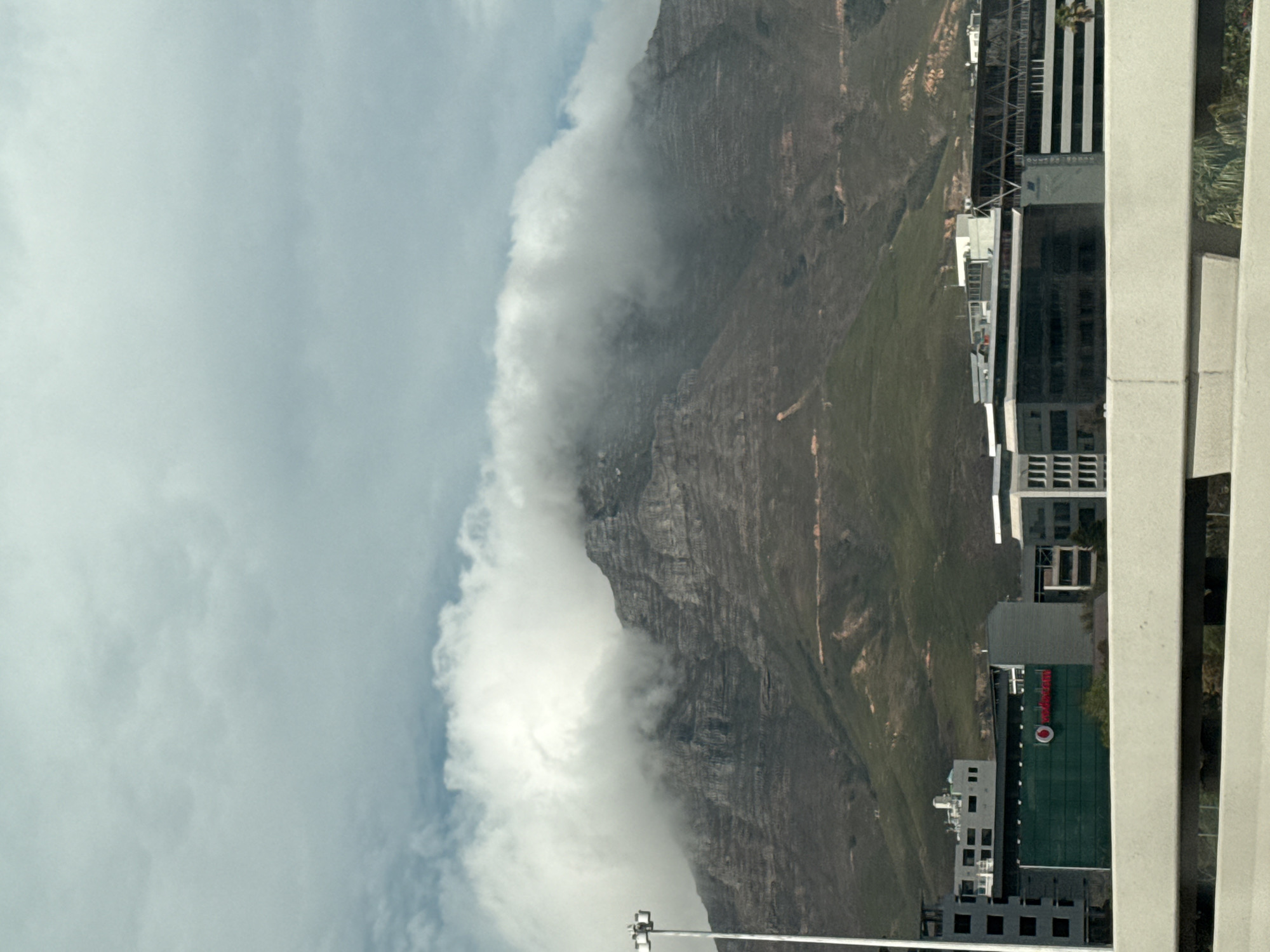 The image depicts Table Mountain in Cape Town, South Africa, partially obscured by clouds. In the foreground are buildings, including one with a red and green logo. The sky is overcast.