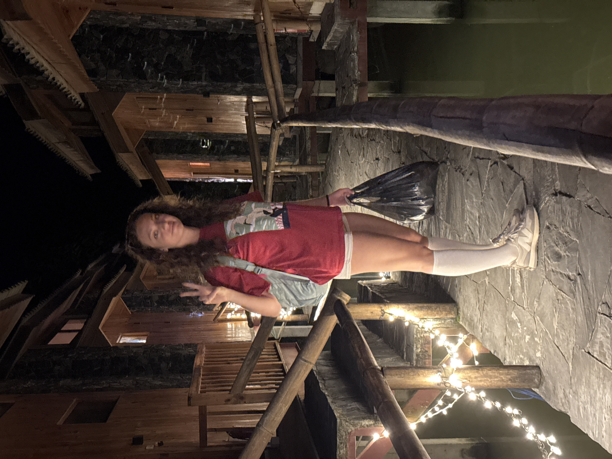 A young woman with curly brown hair is standing outdoors at night in what appears to be Du Già Commune, Vietnam. She is wearing a red t-shirt, light-colored shorts, white knee-high socks, and light-colored sneakers. She is holding a dark object in one hand and making a peace sign with the other. The setting includes wooden structures, bamboo railings, stone pathways, and string lights. Some sort of dark body of water is visible in the background.