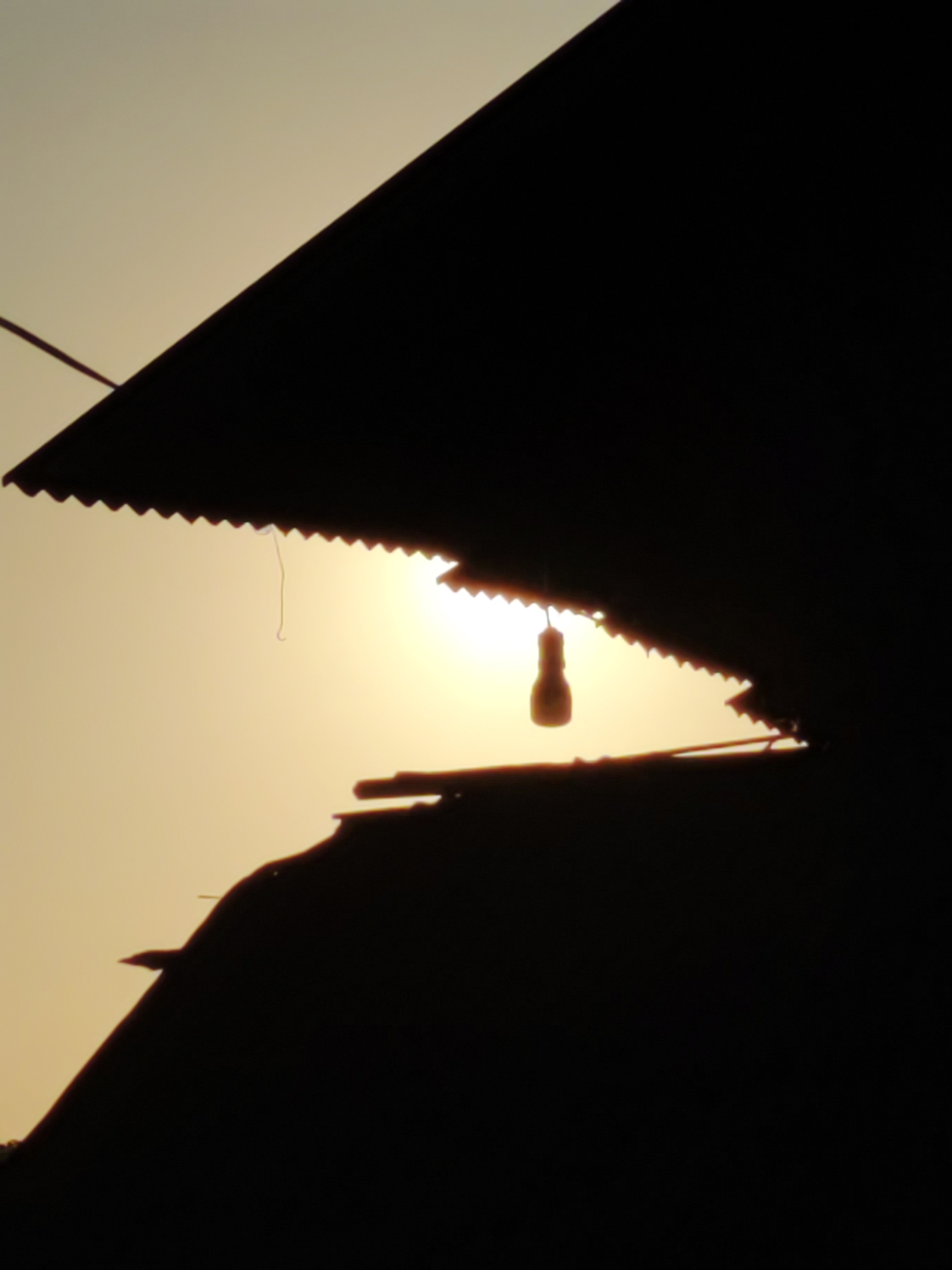 An outdoor, close-up, low-angle silhouette shot captures the sunrise behind corrugated roofing and a single hanging light bulb. The sky is a pale yellow, and the sun is a bright orb peeking through an opening in the layered structures. A thin, dark string hangs from the corrugated roof, attached to a bulb that is a warm, glowing yellow. The silhouette of the roofing creates geometric shapes across the frame, with a jagged edge along the top structure. In the lower portion of the frame, another layer of silhouetted roofing is visible, appearing as a dark, solid mass. The overall impression is one of a rustic, possibly industrial setting at dawn.