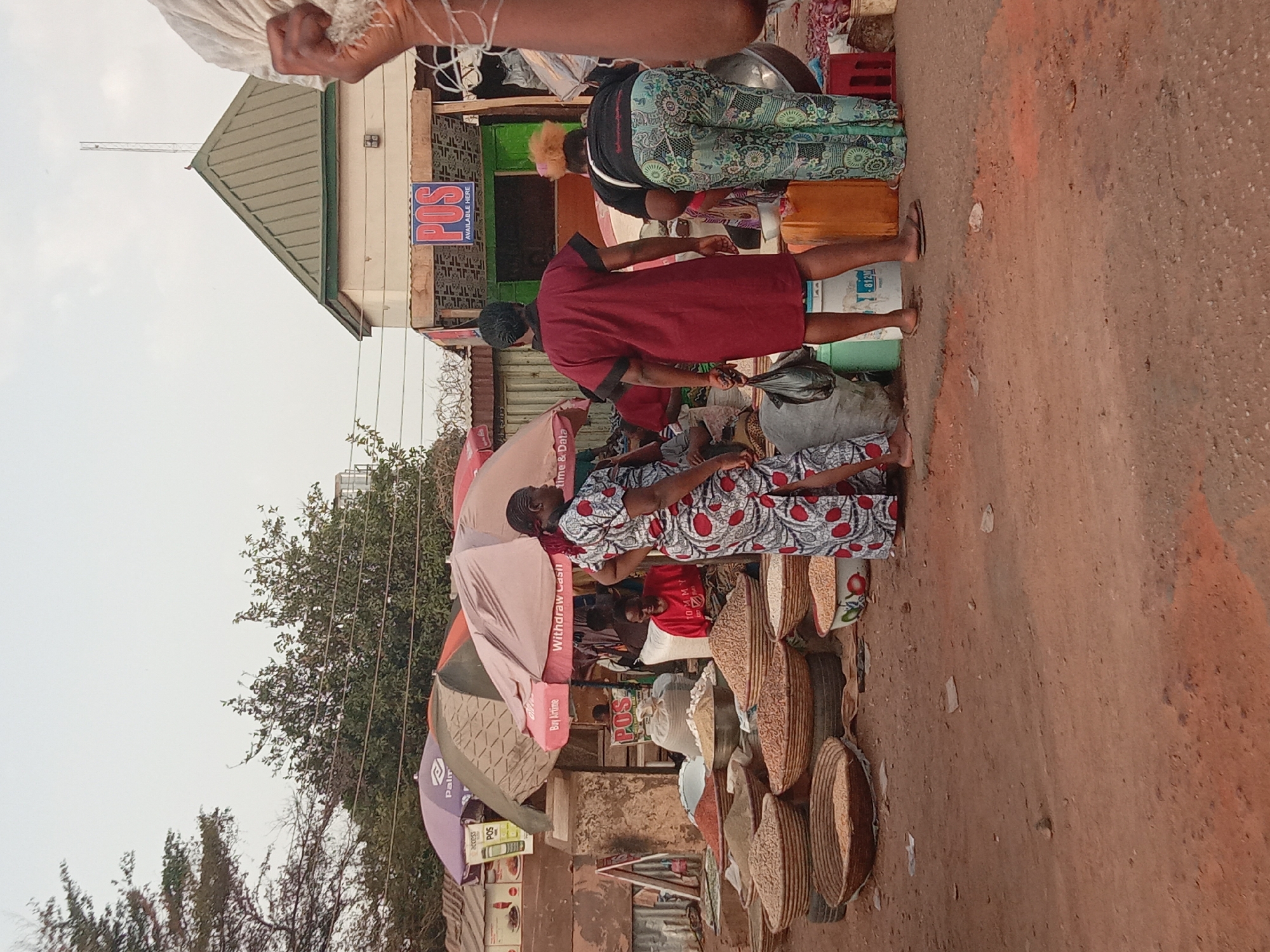 This image shows an outdoor market scene in Kakuri Gwari, Nigeria. Several people are visible, including a woman in a red dress apparently interacting with someone seated amidst a display of woven baskets. Another woman with patterned pants and blonde hair appears to be looking down at the scene. A building with a green roof stands behind the market stalls, and there are signs indicating "POS Available Here", "Withdraw Cash", "Buy Airtime", and "Palir." There are several umbrellas providing shade, and the ground is a reddish-brown color, suggesting a dirt or unpaved road. The presence of market stalls indicates the location is likely a commercial area within the city.