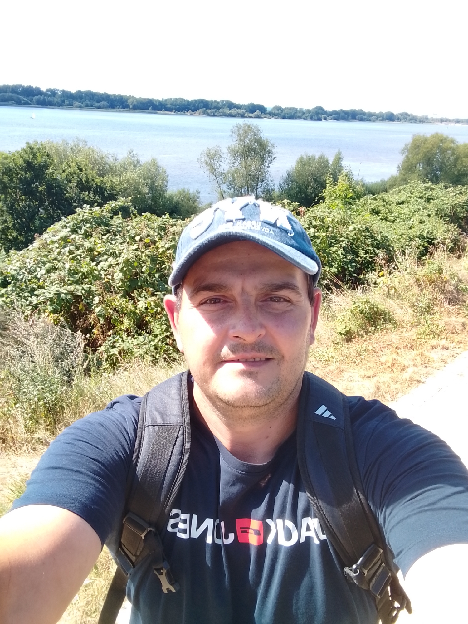 A man is taking a selfie outdoors, wearing a dark blue t-shirt with white lettering and a blue baseball cap. He has a black backpack on. The background shows a body of water, possibly the Elbe river, with trees and vegetation on the shore. The sky is clear and bright. The image appears to have been taken on a sunny day. The man's t-shirt has the brand "JACK & JONES" visible. The scene is likely in the vicinity of Wedel, Germany, a town known for its proximity to the Elbe river.