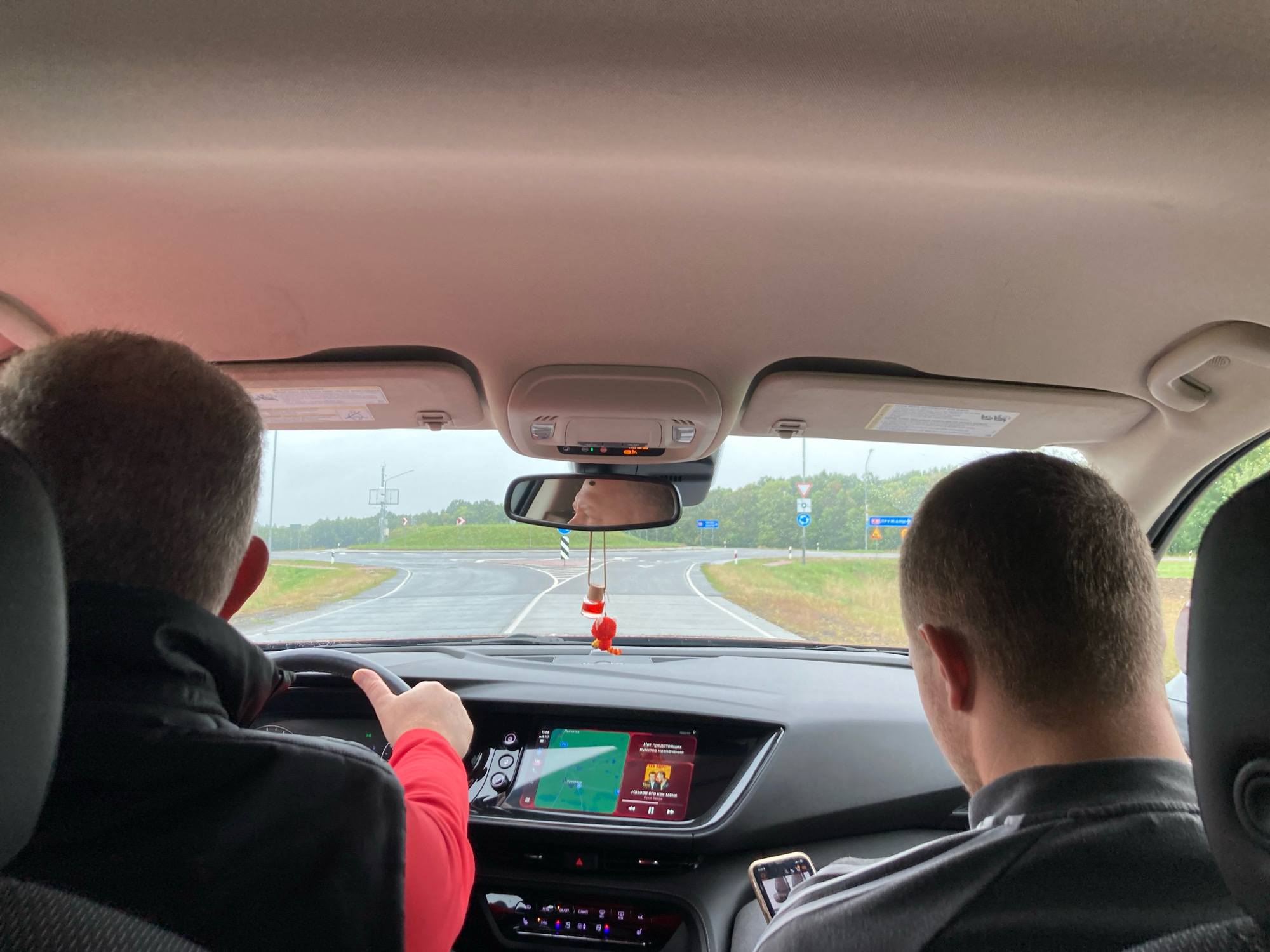 The image shows the interior of a car viewed from the back seats, looking forward. Two men are visible. The man in the driver's seat, wearing a black jacket with a red sleeve, is holding the steering wheel. The second man, in the passenger seat, is looking down at his phone. The car's dashboard displays a navigation system and media player. Outside the car, a road with multiple lanes curves through a landscape of trees and overcast skies. The road signs suggest a location in Агародніцкі сельскі Савет, Belarus.