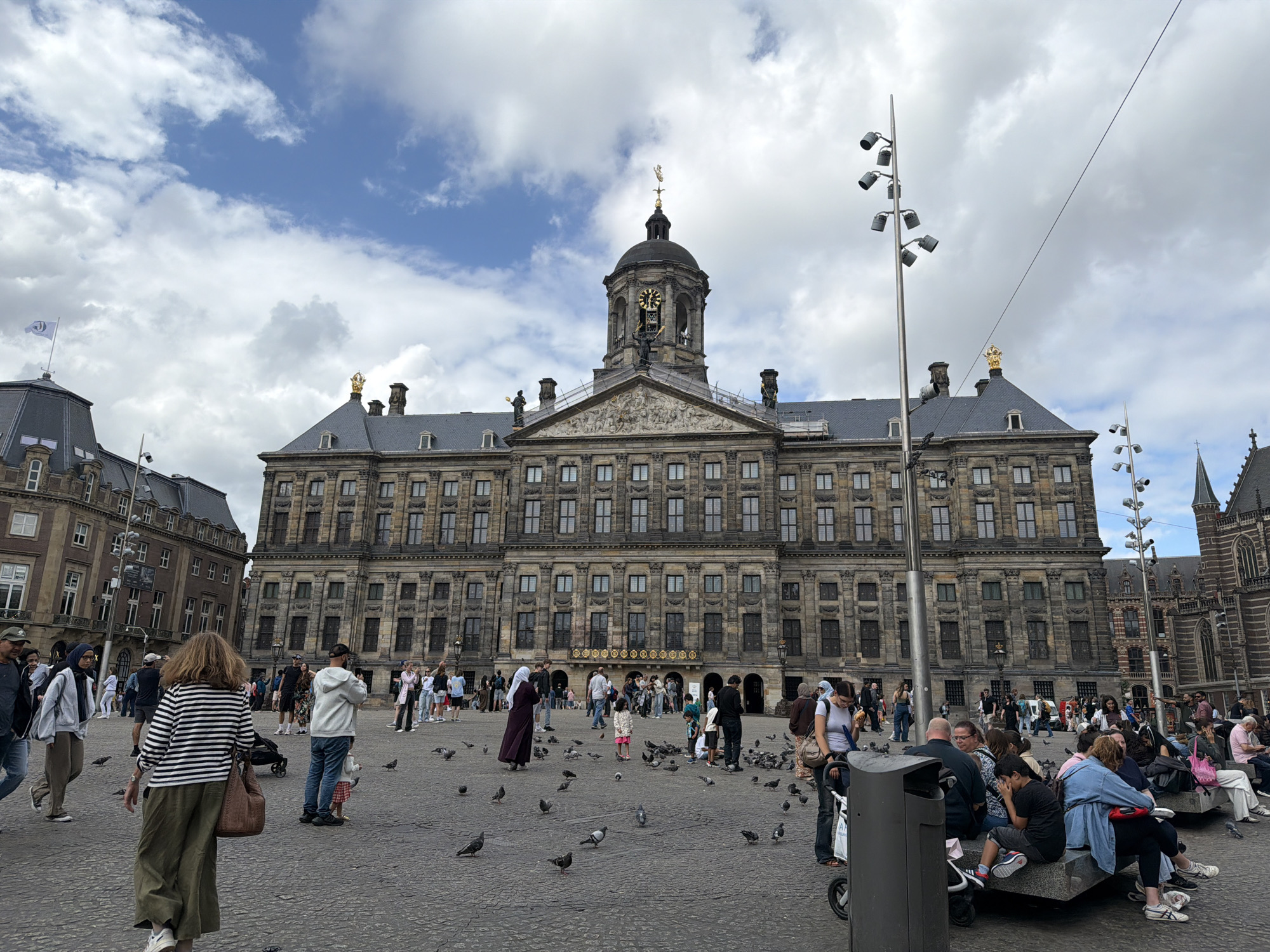The image depicts Dam Square in Amsterdam, Netherlands, on a cloudy day. A large, classical building, identified as the Royal Palace of Amsterdam, dominates the background, its ornate facade featuring numerous windows and decorative elements. A prominent clock tower with a dome rises from the center of the building. In front of the palace, a large, paved plaza is populated with numerous people and pigeons. Various individuals are walking, standing, and sitting in groups on benches, engaged in conversations or observing the surroundings. The people are dressed in casual clothing, indicative of a public gathering or tourist activity. Several tall lamp posts with multiple lights are scattered throughout the plaza. To the left of the palace, another building with a peaked roof and a flag can be seen. The atmosphere appears to be lively yet relaxed, with a mix of locals and tourists.