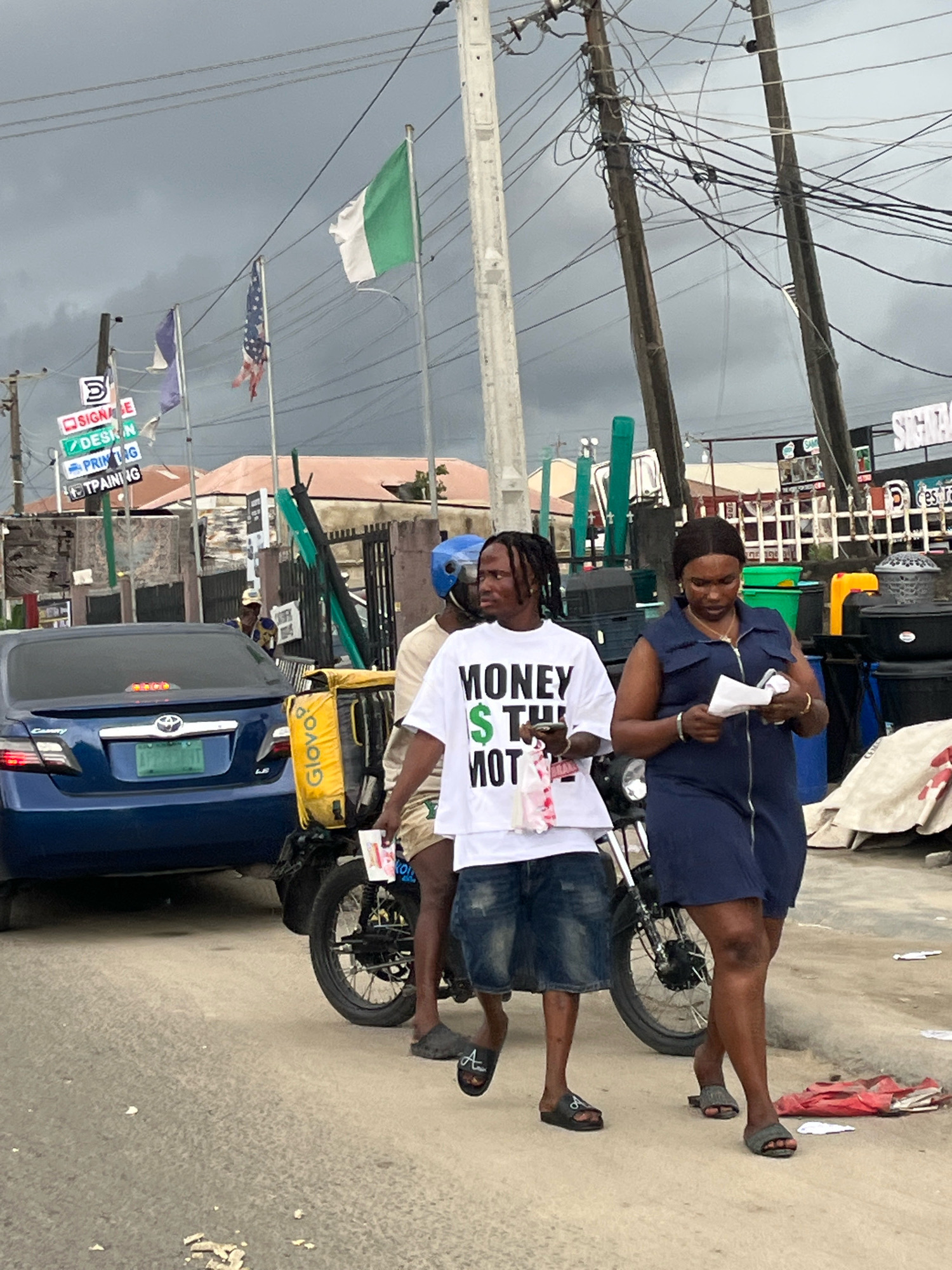 In Ado, Nigeria, a street scene unfolds under a cloudy sky. A blue Toyota Camry is parked on the left, its rear bumper visible. To its right, a motorcycle with a yellow Glovo delivery bag is positioned. Three people are in the foreground. On the left, a young man with braided hair wears a white t-shirt with the text "MONEY IS THE MOTIVATION" printed in green and black. He holds a red and white item in his hand. Beside him, a woman in a navy blue sleeveless dress looks down at some papers she holds. Behind them, another person, partially obscured, wears a light-colored shirt and a blue helmet, seated on the motorcycle. In the background, utility poles with tangled wires dominate the upper portion of the frame. Signs indicating "SIGNAGE," "DESIGN," "PRINTING," and "TRAINING" are visible on the left. The environment appears to be a commercial area with shops and buildings.
