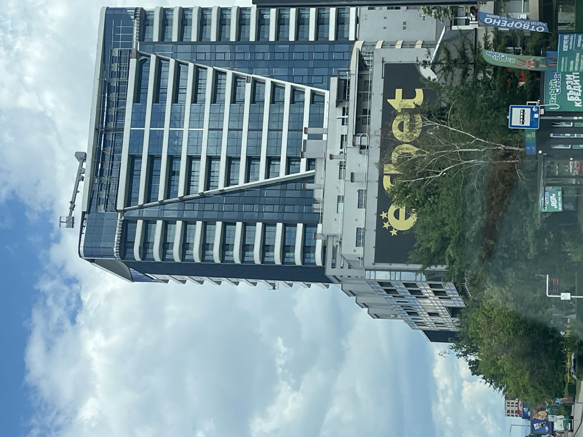 In a cityscape on a cloudy day, a tall, modern building with a glass facade dominates the scene. The building features multiple floors with horizontal white elements and staggered balconies. A large black billboard with the yellow text "e" and stars is prominently displayed on a lower section of the building, partially obscured by trees. To the right of the billboard, a blue banner reads "ОТВОРЕНО". In the foreground, green trees and foliage are visible, along with signs and structures that suggest a street or public area. A bus stop sign is present. The overall impression is of an urban environment with contemporary architecture.