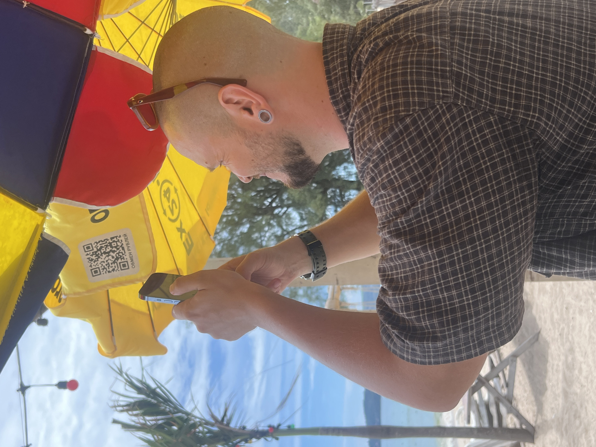 In Choeng Thale, Thailand, a bald man with a goatee and ear gauges is holding a smartphone to take a picture. He is wearing a plaid shirt and sunglasses are perched on his head. He is standing under a multi-colored umbrella, possibly at a beachside establishment, as evidenced by the palm tree, sand, and ocean in the background. A QR code with Russian text is visible on the umbrella.