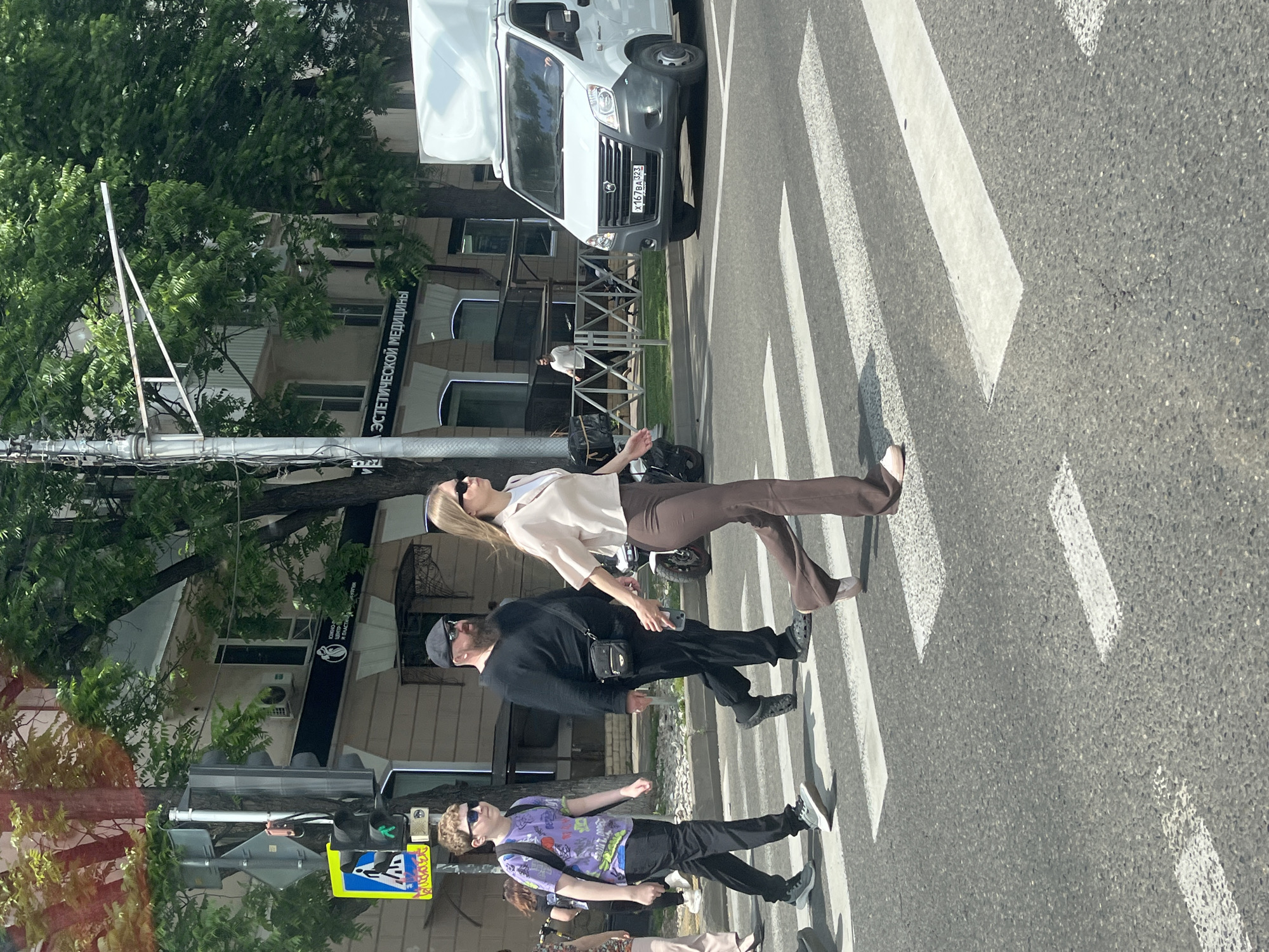 The image shows a group of people crossing a street at a pedestrian crossing in Krasnodar, Russia. In the foreground, a young boy with blond hair, wearing a purple t-shirt and black pants, is walking across the street. To his right, a man with a beard and a black cap, wearing a black sweatshirt and black pants, is also crossing. A woman in a light-colored blouse and brown pants is walking in front of them. Behind them, a white van is stopped at the intersection. In the background, there are buildings and trees lining the street. A traffic light is visible on the left side of the image, with a pedestrian crossing signal illuminated. The scene appears to be taking place on a sunny day.