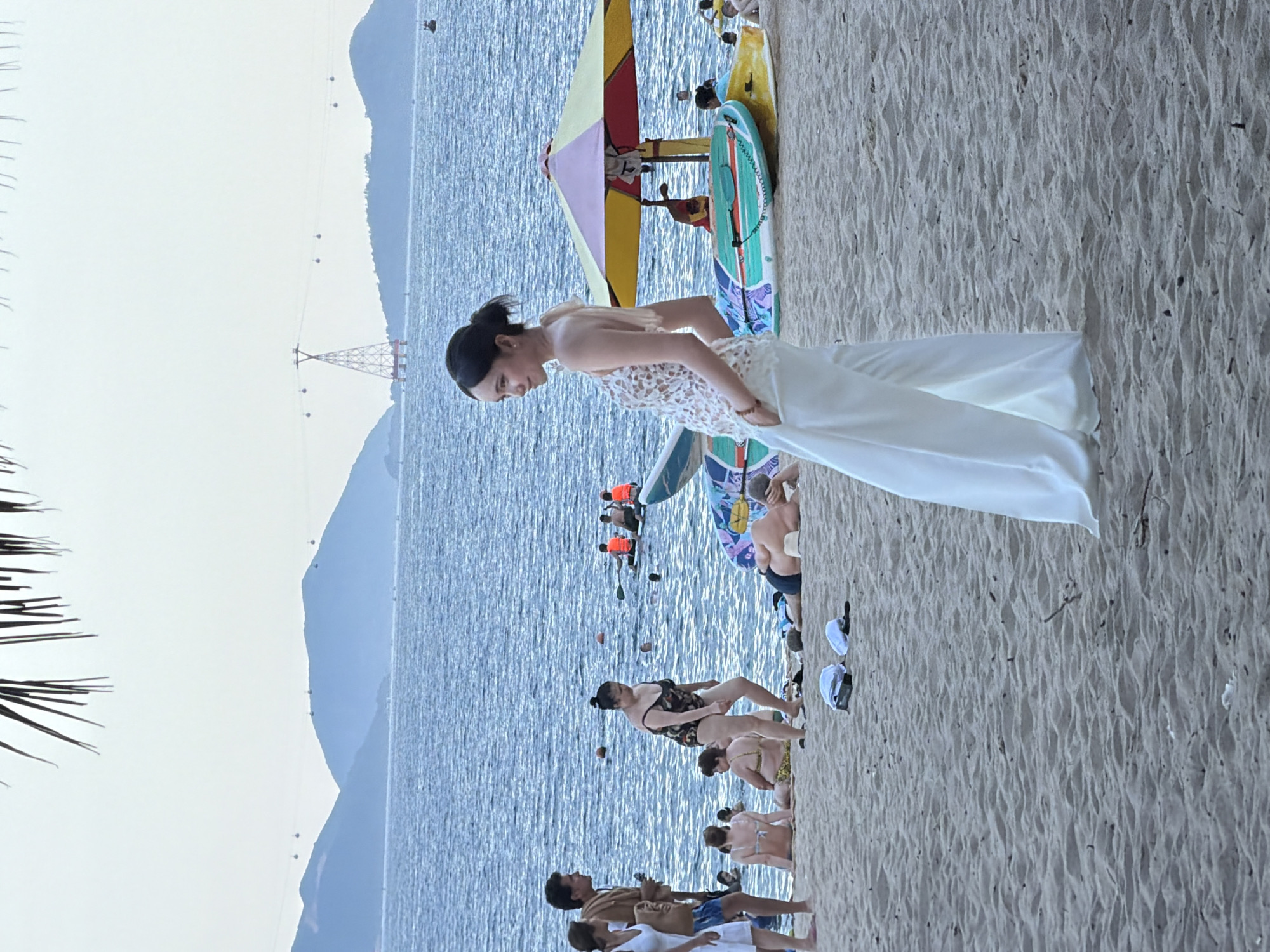 The image depicts a beach scene in Nha Trang, Vietnam, with the Nha Trang Cable Car system visible in the background, spanning across mountains and the sea. In the foreground, a woman in a white lace top and wide-leg pants stands on the sand, facing left with her hands in her pockets. Behind her, the beach is populated with several individuals. Some are in the water, including people on paddleboards and others swimming. Beach umbrellas and surfboards are also present. The overall atmosphere suggests a relaxed day at the beach.