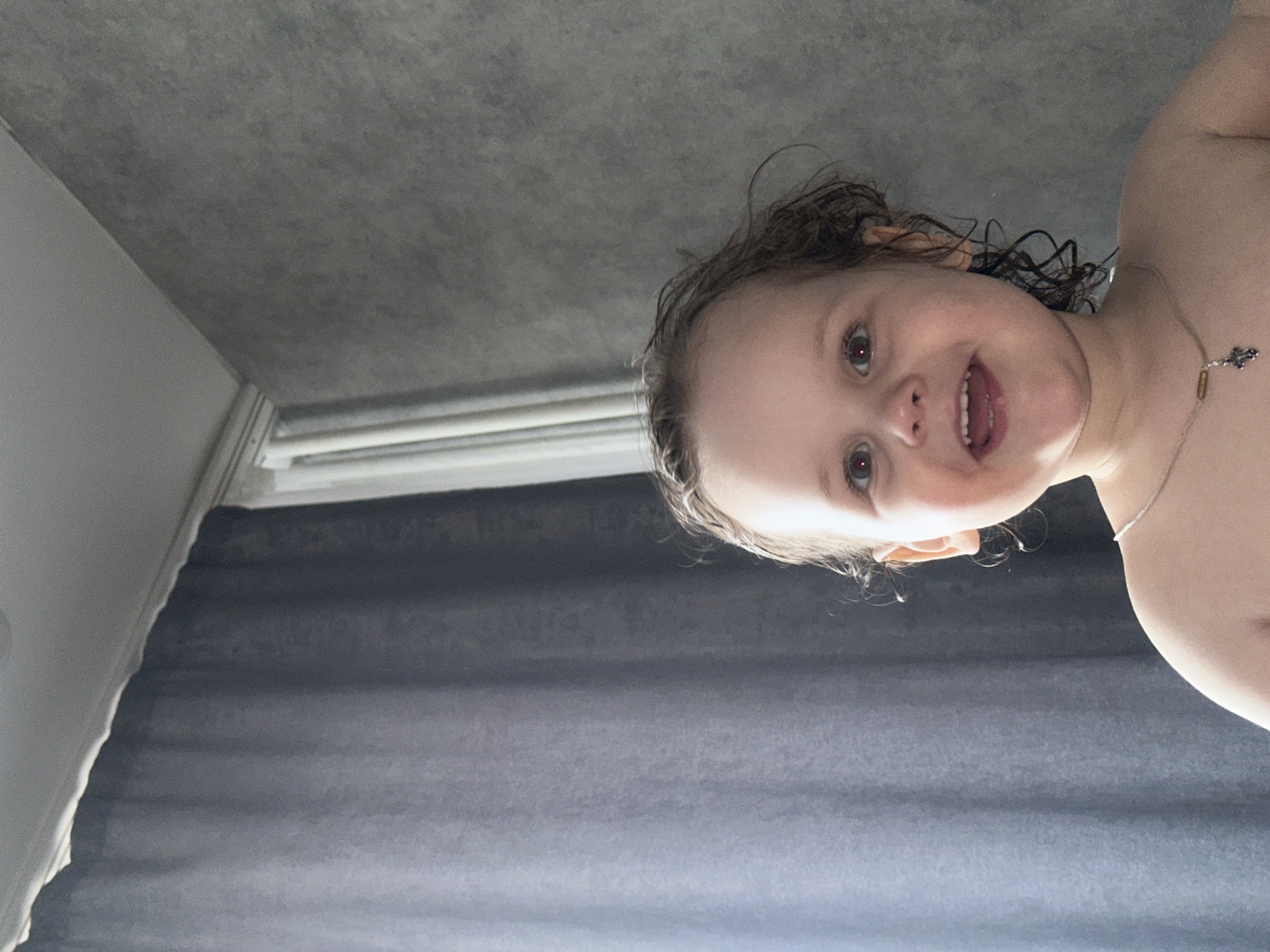 A young child is looking down at the camera. The child has wet hair and is wearing a necklace. In the background, there is a textured gray wall and a window with gray curtains. There is no information in the image to suggest it was taken in Chaikovsky, Russia.