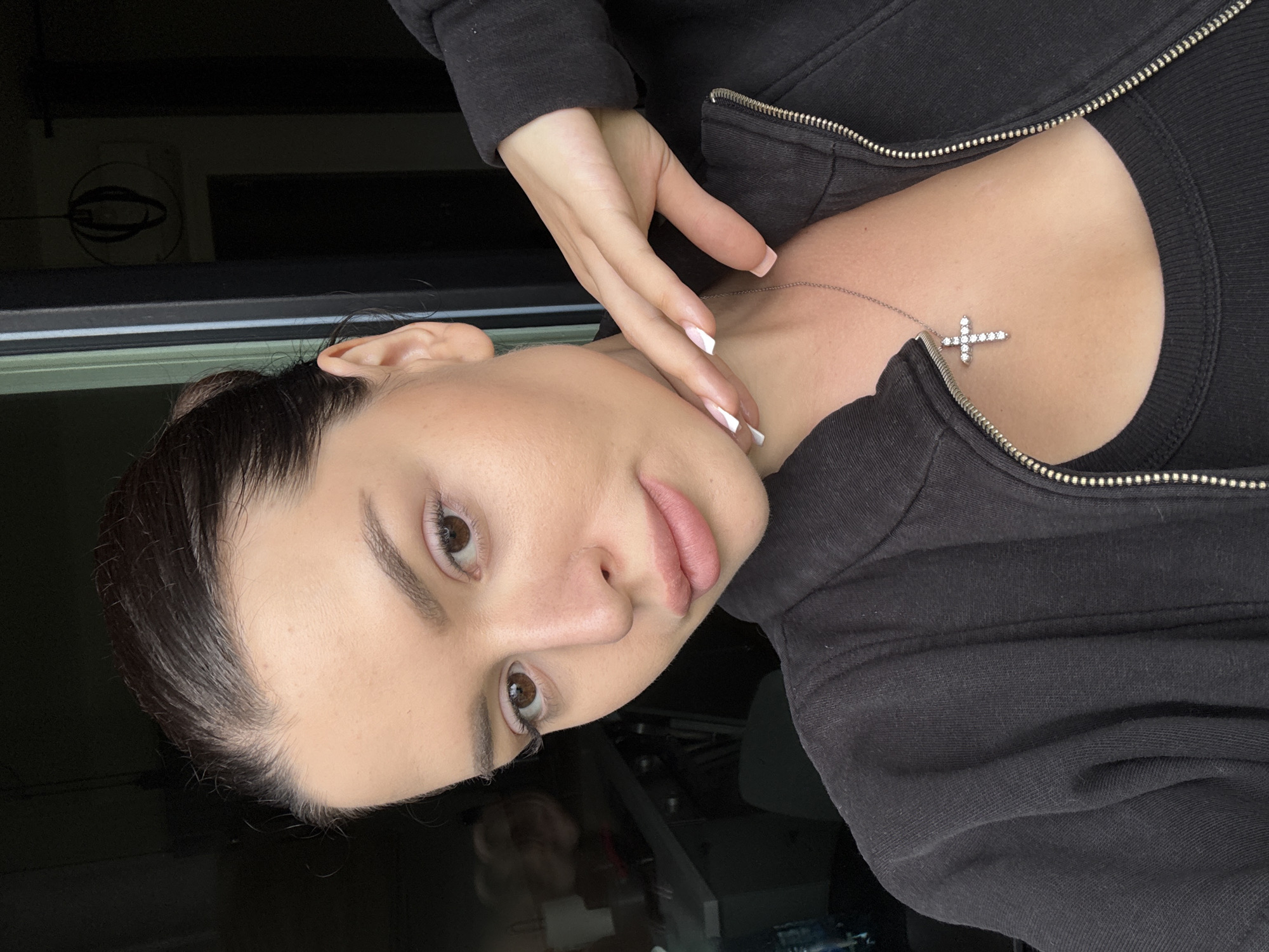 A close-up, vertical, indoor shot features a young woman as the sole subject. Her head is tilted, and her gaze is directed towards the camera. She has dark hair pulled back, revealing her face, which is illuminated by soft lighting. She wears a black zip-up hoodie over a black tank top. A silver necklace with a small, diamond-encrusted cross pendant rests on her collarbone. Her right hand, with neatly manicured, pale pink nails, is gently placed on her neck and décolletage, with her fingers delicately touching her chin. The background is dark and indistinct, with some subtle reflections suggesting an indoor environment, possibly near a window. The overall impression is one of a candid selfie, with a casual and intimate feel. There is no visible text in the image. The time of day and weather are not discernible from the image.