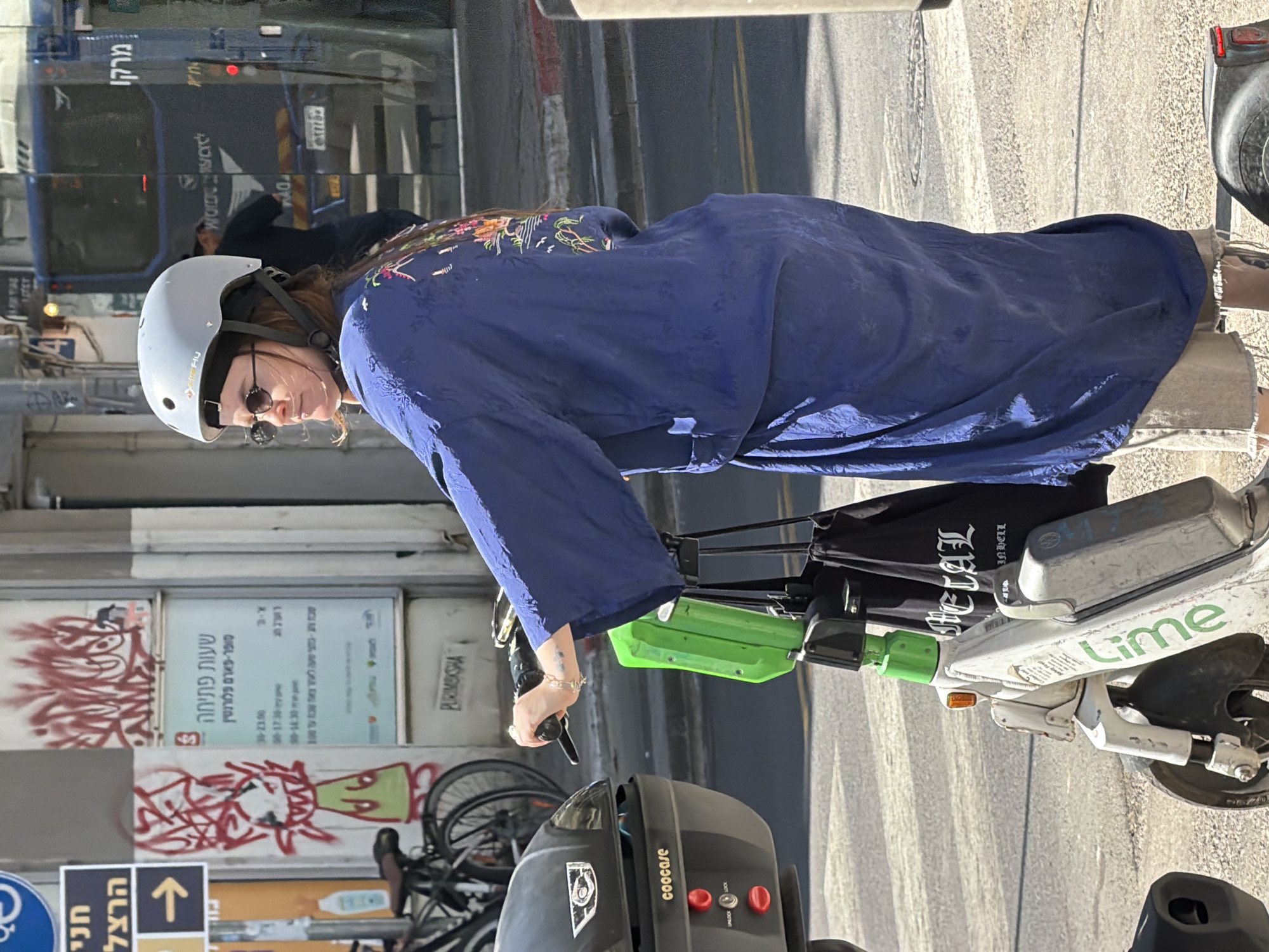 The image depicts a person riding a Lime electric scooter on a Tel Aviv street. The rider, wearing a white helmet, round sunglasses, and a loose, blue, embroidered top, is situated on a sidewalk near a building with graffiti and store signage in Hebrew, including a sign indicating it is the Super-Pharm Florentin. The street is also visible, with a bus parked nearby. The Lime scooter features a black Coocase storage box on the rear, and a black bag hangs from the handlebars with the word "HELAL" printed on it.