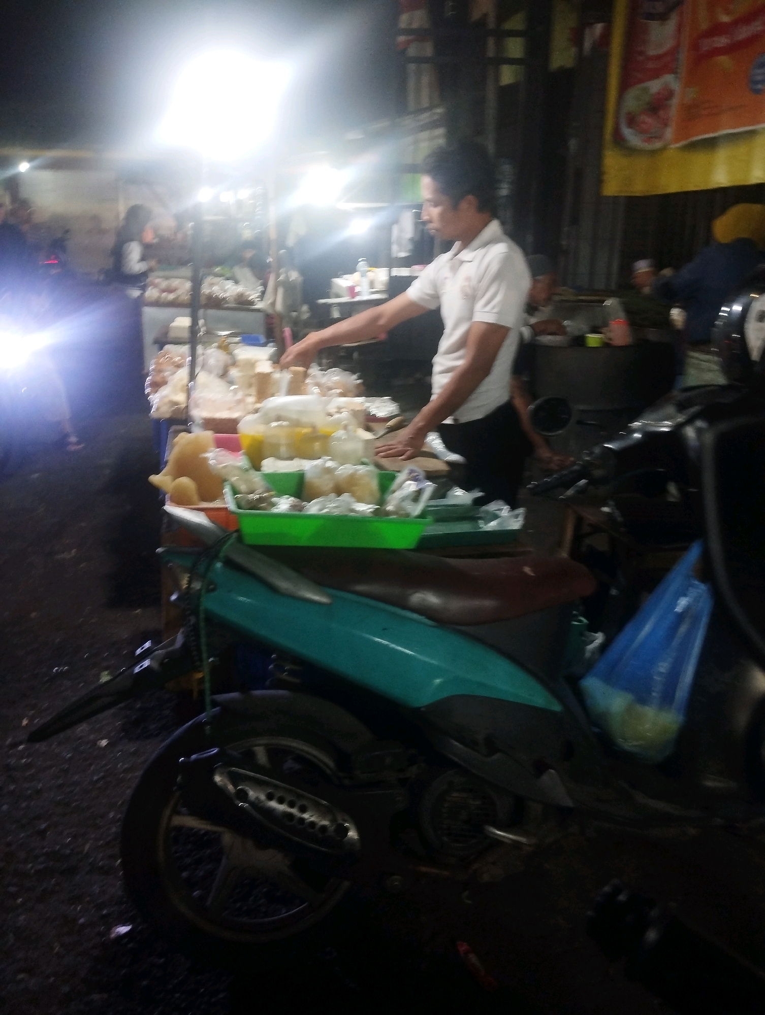 An outdoor scene, likely a street food stall at night, is depicted in the city of Malang, Indonesia. A man in a white polo shirt is actively engaged at a stall filled with various food items, some of which appear to be wrapped in plastic or contained in trays. His right hand is reaching towards the food. A motorcycle, teal and black in color, is parked in the foreground, partially obscuring the stall and the ground which is covered in dark asphalt. A blue plastic bag containing light-colored items is hanging from the motorcycle. In the background, other individuals are present, and a bright light source, possibly a street lamp or stall lighting, creates a strong glare. Yellow and orange banners are visible further in the background, suggesting a commercial area.