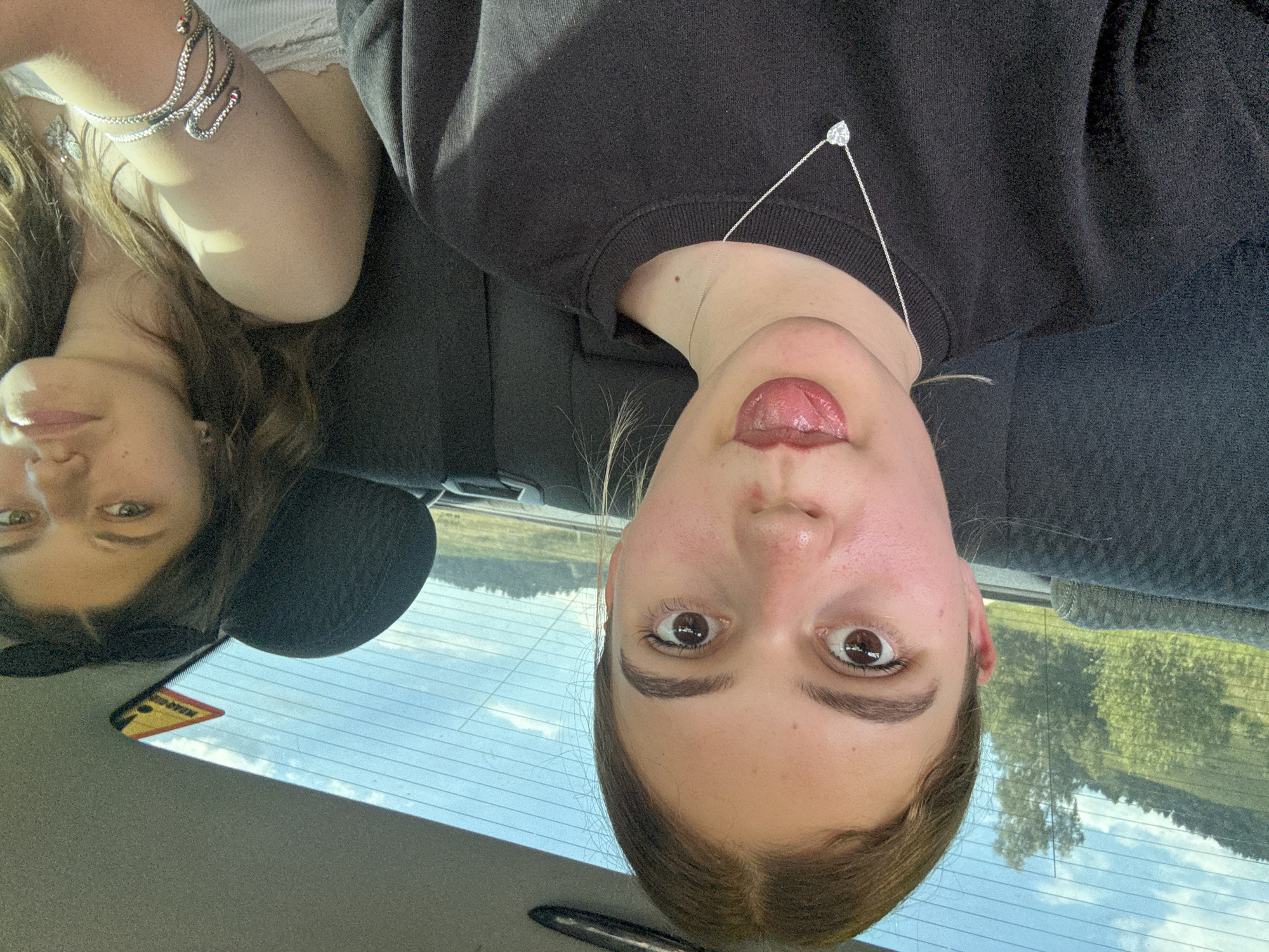 The media file shows a selfie taken by two young women inside a car. One woman is in the foreground, upside down, with her face close to the camera. She has brown eyes and is sticking her tongue out, wearing a black sweatshirt and a heart-shaped pendant necklace. The other woman is partially visible in the background, also upside down, wearing a white tank top and a snake-like bracelet with red accents. She has long brown hair and is looking towards the camera.
The scene appears to be set during the daytime, with sunlight filtering into the car. The background visible through the car windows shows a landscape with trees and a blue sky with clouds, suggesting an outdoor setting. A triangular sticker with yellow and black warning symbols is visible on the rear window, indicating a possible hazard or information.
The overall mood seems playful and casual, indicated by the woman's expression. The image is a snapshot of a moment, likely during a road trip, with the subjects enjoying their time together. There is no other visible text in the image besides the warning sticker.