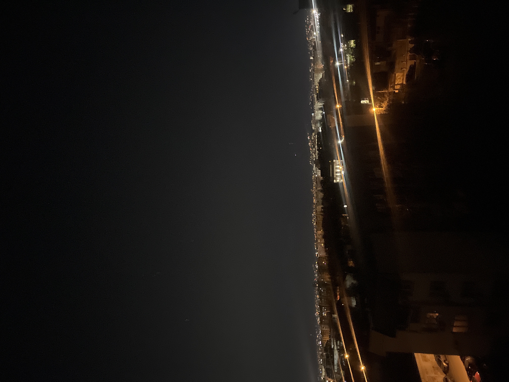 This is a nighttime view of a city along a coastline. The left side of the image is predominantly a dark body of water, possibly the Black Sea. The right side shows a city, identified by the presence of streetlights and building lights. Although the specific location in Sofia, Bulgaria is unknown, the image depicts an aerial view from a high vantage point of a well-lit urban area with a coastline adjacent to a dark body of water.