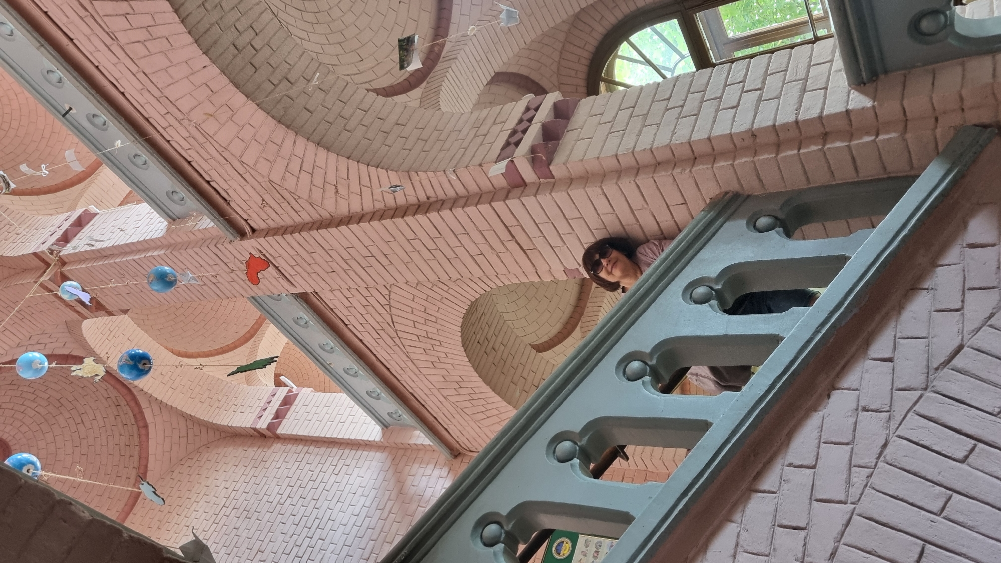 A woman wearing sunglasses and a pink shirt leans against a pink brick wall in a grand staircase, possibly within the Chernivtsi National University complex in Chernivtsi, Ukraine. She is positioned behind a light blue ornate railing. The architecture features arched ceilings and decorative brickwork. Several blue globe-shaped ornaments and various cutout figures hang from strings suspended from the ceiling, adding a whimsical element to the scene. The lighting suggests it is daytime, with natural light filtering in from a window in the background.