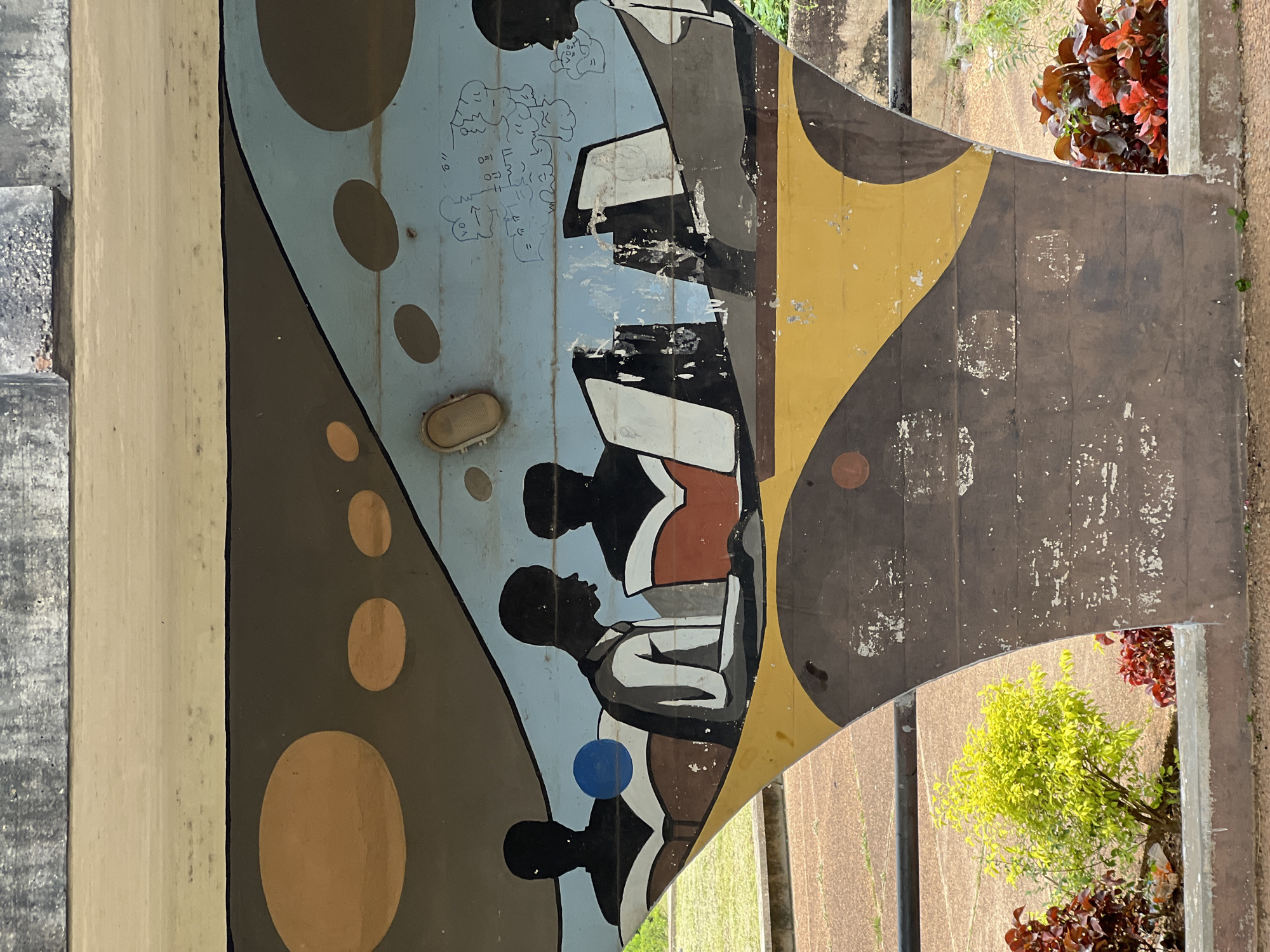 This is a medium shot, eye-level photograph of a mural painted on a concrete structure. The mural depicts several silhouetted figures of people, appearing to be students, engaged in reading. They are seated, with open books in front of them, and the composition suggests a group of individuals learning together. The mural uses a limited color palette of browns, yellows, blues, and blacks, with some white accents on the figures and their clothing. The background of the mural features abstract shapes and some crudely drawn figures and text, possibly representing thought bubbles or an academic environment. Below the mural, there are planters with vibrant green and red foliage, and in the background, a grassy area and a dirt path are visible. The concrete structure itself shows signs of age and wear, with peeling paint and discoloration. This mural is located at the Obafemi Awolowo University (OAU) in Ile-Ife, Nigeria, specifically on the underside of an overpass bridge that leads into the university campus.