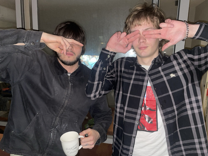 Two young adult males are depicted indoors, posing with their hands covering their eyes. The specific location within Tel-Aviv, Israel, is not discernible from the image, but the setting appears to be a residential interior, possibly near a window or glass door.
The subject on the left, with dark hair and a beard, wears a dark, possibly black, zippered work jacket or chore coat, which appears stained or dusty with light-colored marks. His right hand covers his eyes, with fingers spread across his forehead. His left hand holds a white ceramic mug with dark text that reads "Happiness is not about having everything you want, but about enjoying everything you have." A small tattoo is visible on his left hand.
The subject on the right, with blonde or light brown shaggy hair, wears an unbuttoned black and grey plaid long-sleeve shirt over a white t-shirt featuring a red graphic design. His left wrist displays a metallic chain bracelet. Both of his hands are positioned over his eyes, forming "V" shapes with his fingers.
The background is dimly lit and out of focus, showing indistinct elements consistent with an indoor environment. The subjects are illuminated by a direct light source, suggesting an artificial light or camera flash.