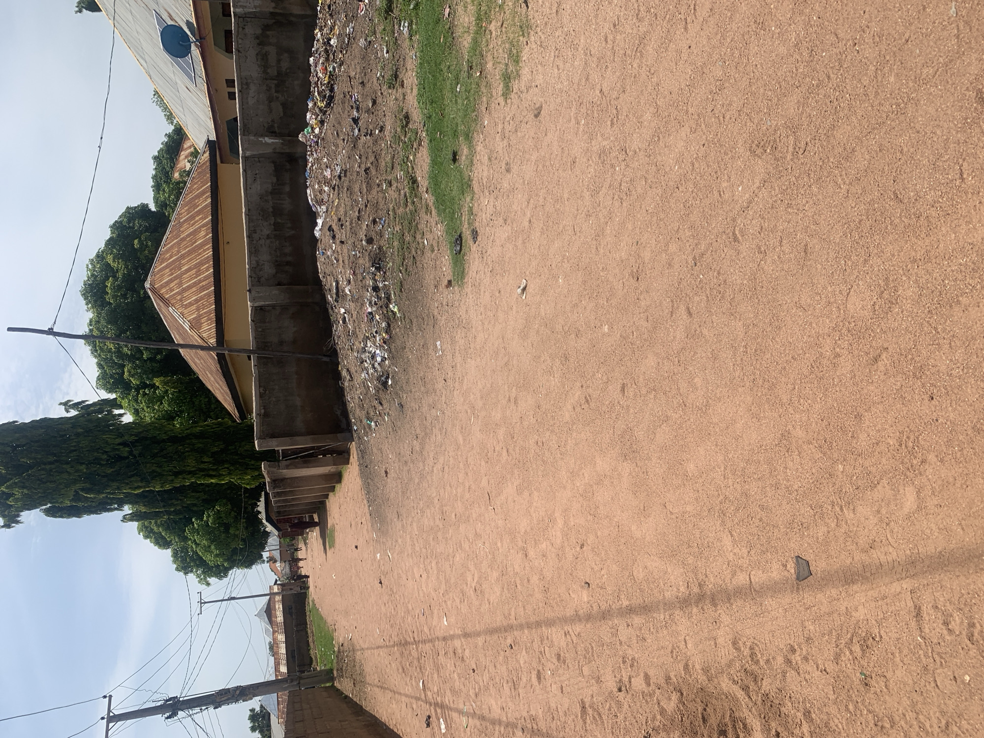 The image shows a dirt road in the city of Jauro Yinu, Nigeria, lined with concrete walls and houses with corrugated metal roofs. A large pile of garbage is visible on the right side of the road, and a few people are walking in the distance. The sky is clear and blue, with a few clouds. The road appears to be a well-trafficked area, with tire tracks and footprints visible in the dirt. The presence of a satellite dish on one of the roofs suggests that modern amenities are available in the area.