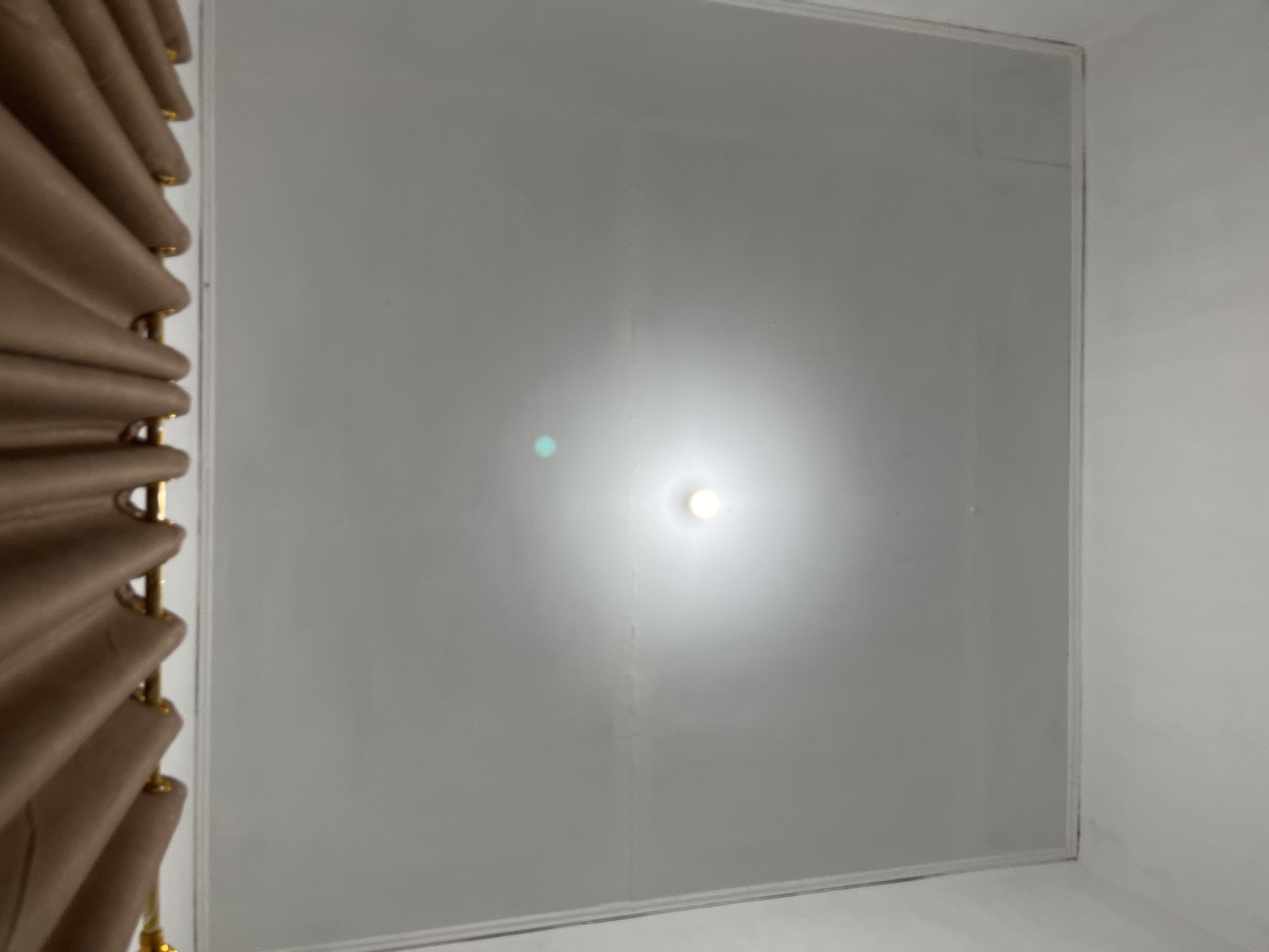 The image displays an upward view of a white ceiling in an indoor room, located in Lhokseumawe, Indonesia. A single, bright light source, appearing as a circular white glow, is prominently featured near the center of the ceiling. A faint greenish-blue lens flare is visible to the left of the light.
The ceiling itself is composed of light grey or white panels, outlined by subtle darker lines, suggesting a modular or tiled construction. Some faint vertical smudges or streaks are visible on the ceiling surface, particularly below the light fixture.
Along the left edge of the frame, a segment of a light brown or beige curtain or blind is visible. It features soft, wavy folds or pleats running vertically, with small golden, possibly metallic, rings or fasteners interspersed along its length. The curtain extends vertically across the frame's left side, partially framing the ceiling. The surrounding walls are also visible in the corners, appearing white. No people or activities are depicted, and there is no visible text. The scene is illuminated by the artificial light, providing a bright, uniform lighting effect.