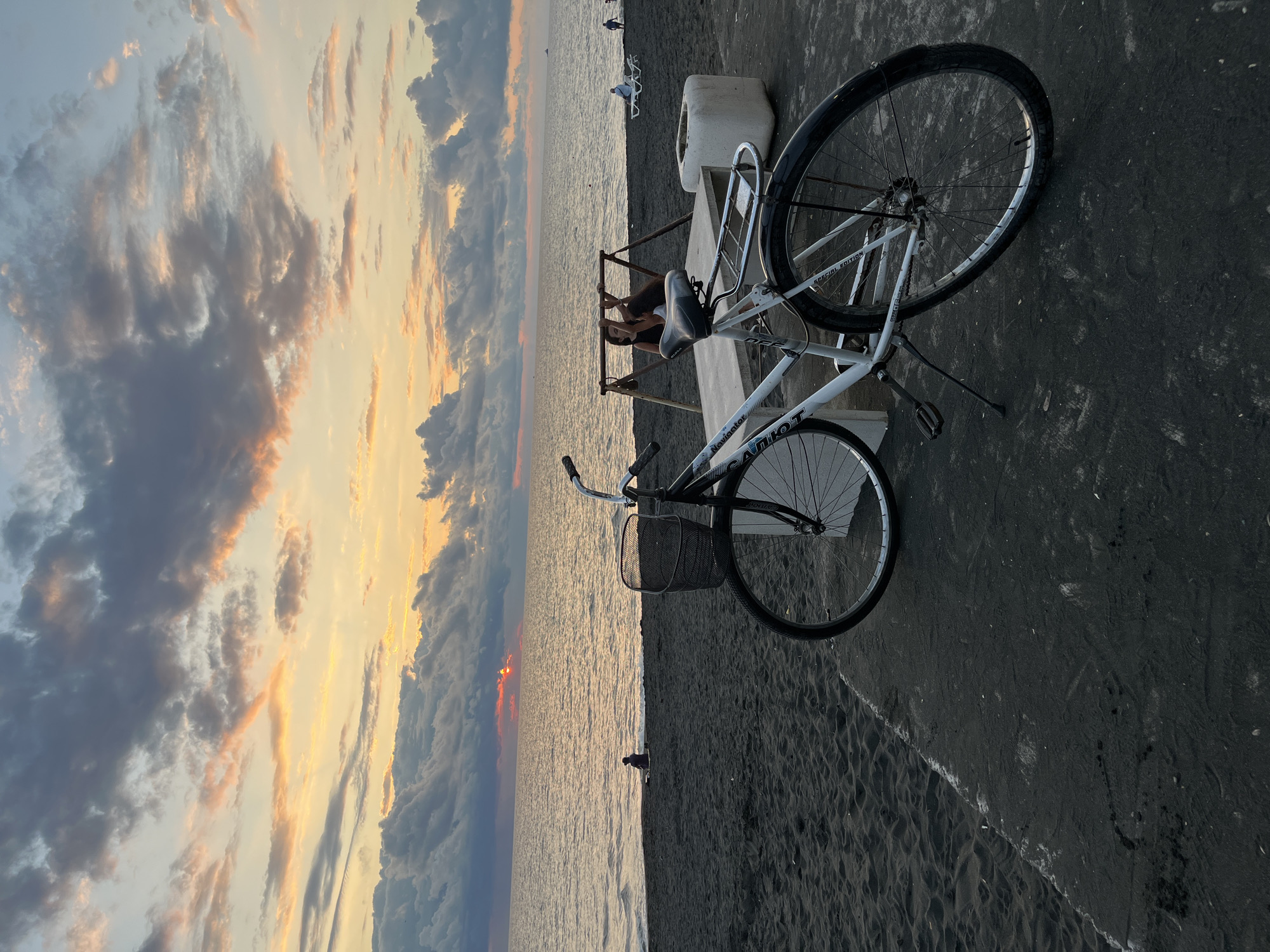 A white bicycle with a basket is parked on a concrete path next to a black sand beach. In the background, the sun is setting over the Black Sea, casting a warm glow on the sky filled with dramatic clouds. Several people are scattered along the beach, some sitting and others lounging on chairs. A swing set with two individuals is visible near the bicycle. The overall scene is one of a peaceful evening by the sea.