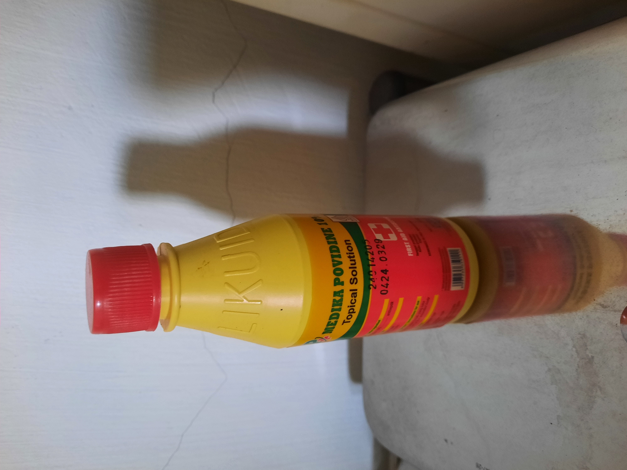 A yellow plastic bottle with a red cap containing "MEDIKA POVIDINE 10% Topical Solution" stands on a shiny, reflective surface. The bottle label is predominantly red with white and green text. A barcode is visible on the lower part of the label. A shadow of the bottle is cast on a light-colored wall behind it. The overall scene suggests a product photograph or an inventory image. The specific location in Lhokseumawe, Indonesia, is not discernible from the visual information provided.