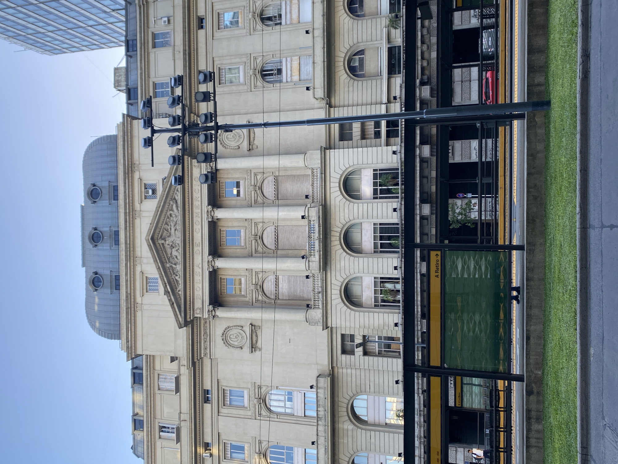 The image depicts a street view in Buenos Aires, Argentina. A substantial, multi-story building with neoclassical architectural elements, including columns, arched windows, and ornate detailing, dominates the left side of the frame. A dome-like structure is visible atop the building. Lettering reading "BOLSA DE COMERCIO" is visible on the building's facade. To the right is a metro stop, indicated by the signs "A Retiro" and the green tiling. There are figures moving near the metro stop, including someone wearing a white shirt. A grassy strip separates the metro from the road, which forms the right edge of the image. A street lamp is visible in the foreground. The sky is light blue.