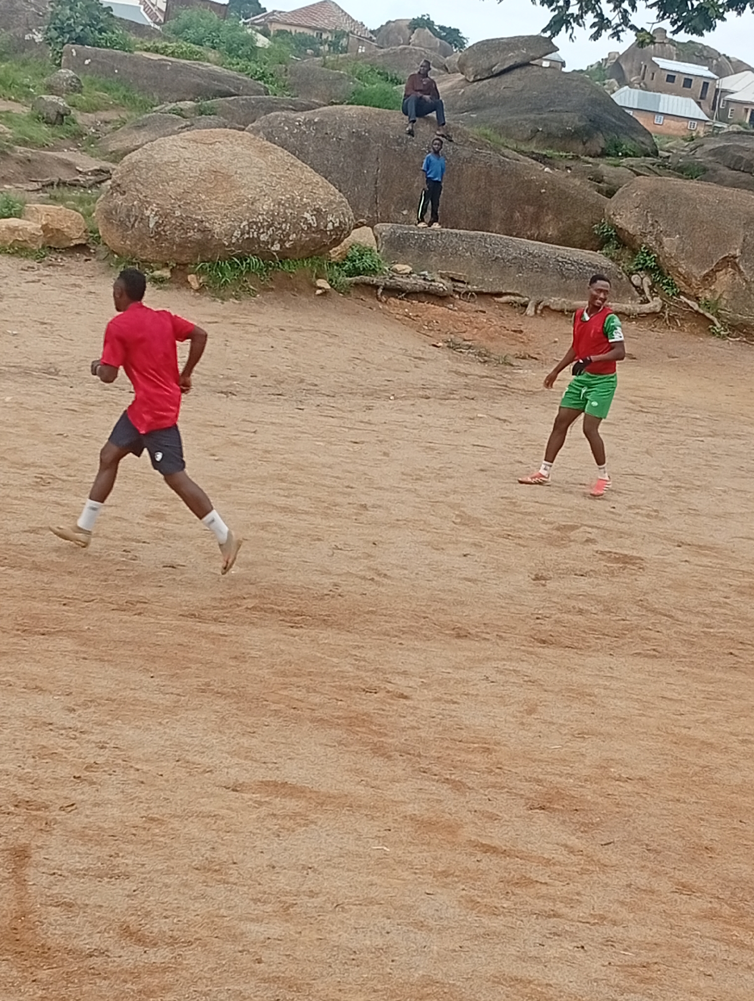 In the city of Unknown, Unknown, two young men are engaged in physical activity on a sandy, uneven terrain. One man, on the left, is wearing a red t-shirt and dark shorts, appearing to be mid-stride as if running or jogging. He has dark hair and is wearing white socks and light-colored shoes. To the right, another man is wearing a red vest over a green shirt and green shorts. He is also wearing white socks and pink athletic shoes, and is looking to his left. In the background, large, weathered rocks dominate the landscape, with some vegetation growing among them. Buildings with tiled roofs are visible on the higher ground behind the rocks. The ground is dusty and appears to be a mixture of sand and dirt, with some small pebbles scattered about. The overall scene suggests an outdoor recreational area in a hilly, possibly arid environment.