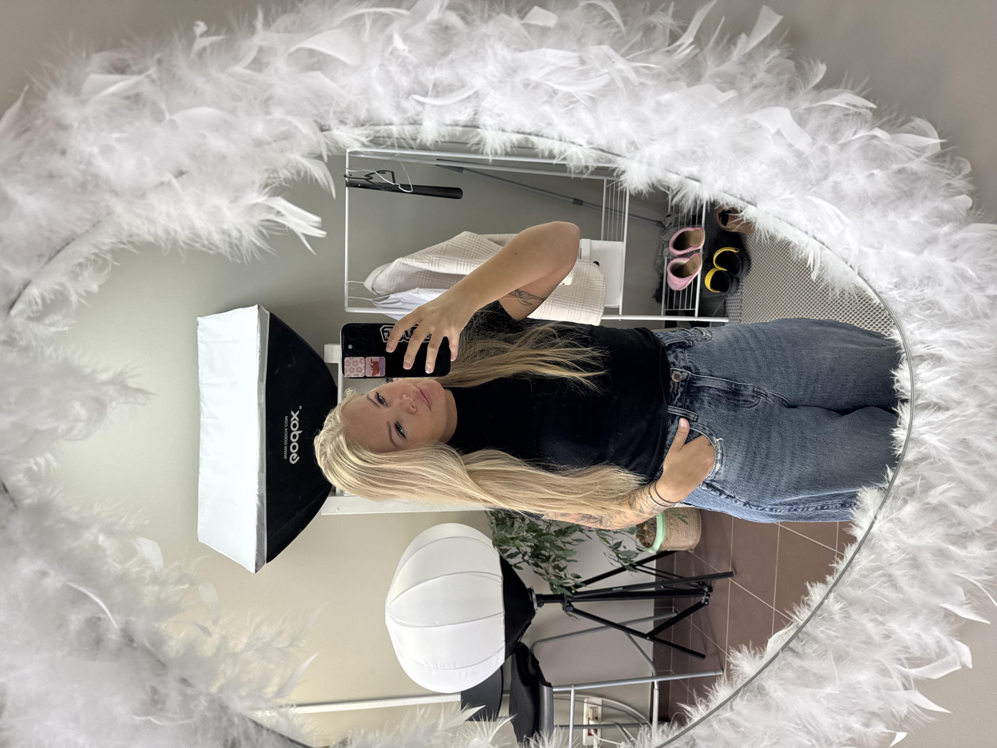 The image depicts a woman taking a selfie in a mirror adorned with white feathers. The mirror reflects a portion of a room with a tiled floor, a plant on a stand, a softbox lighting setup, and a shelving unit holding clothing and shoes. The woman is wearing a black long-sleeved shirt and blue jeans, holding her phone up to capture the image. Her hair is long, blonde, and flowing. Tattoos are visible on her arms. The city and specific location are not discernible from the image.
