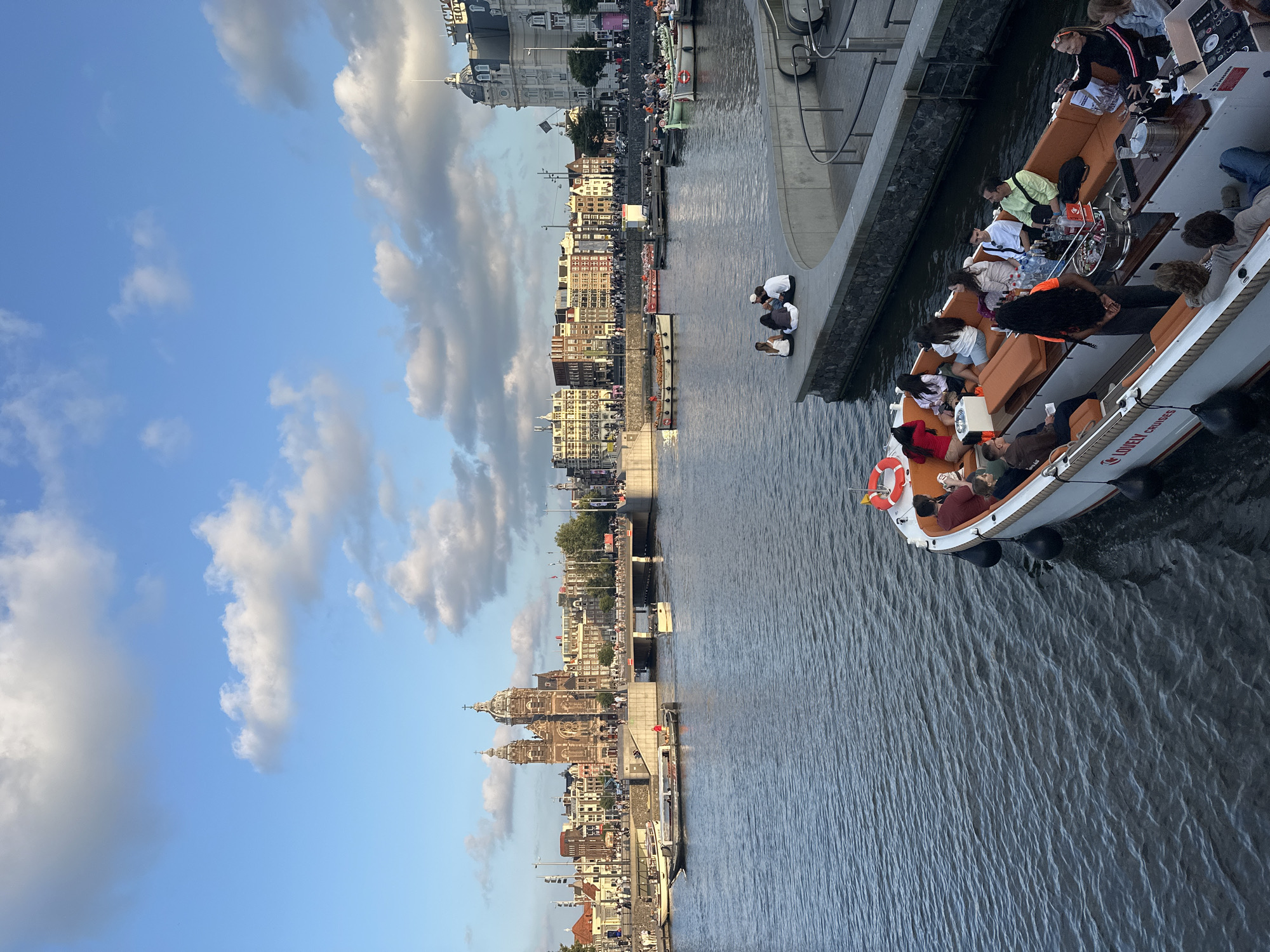 The image shows a canal in Amsterdam, Netherlands, with a boat of tourists in the foreground and a cityscape in the background. The boat is white with orange seating and is filled with people enjoying a canal cruise. The cityscape features a large, ornate church with two towers, typical of Amsterdam's architecture, as well as other historic buildings lining the canal. Several other boats are visible on the canal, and a few people are sitting on the edge of the canal in the middle ground, observing the scene. The sky is blue with scattered white clouds.