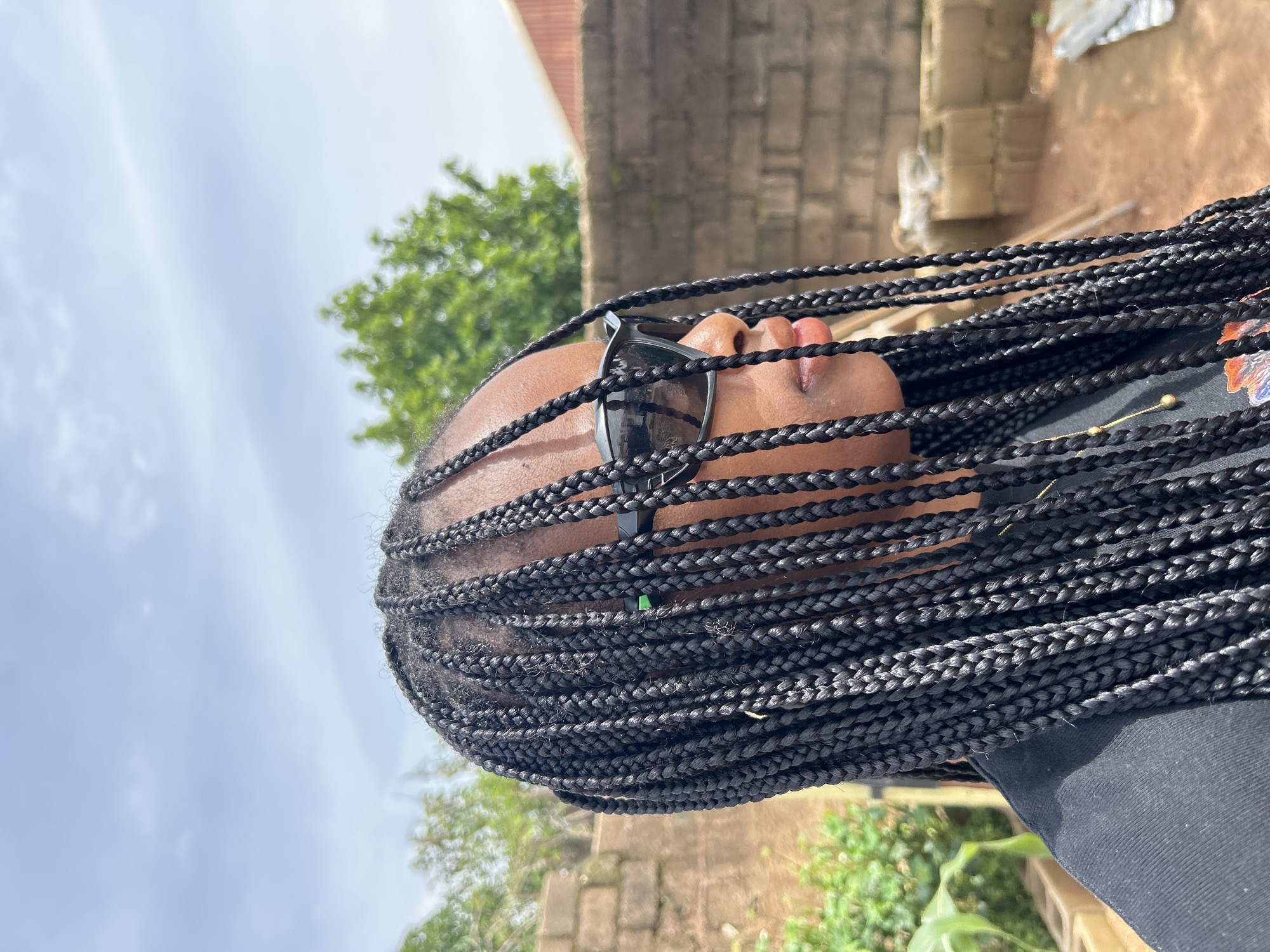 A young African woman with long black box braids is pictured from the shoulders up, in profile, facing right. She is wearing black-framed sunglasses and a black t-shirt. The background appears to be a rural or semi-urban outdoor setting, possibly a courtyard, with a brick wall to the right, some greenery and trees behind, and a cloudy sky overhead. The lighting suggests daytime. The context of Jos, Nigeria is noted, however, no specific landmarks or identifiers within the image confirm this location.
