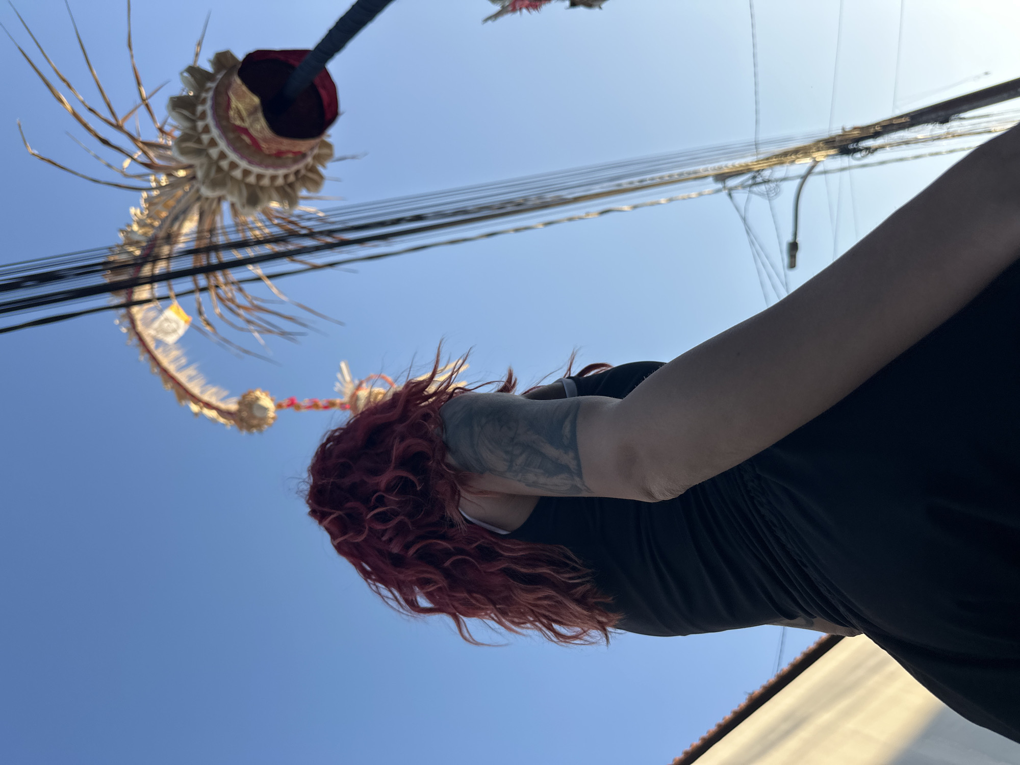 An individual with vibrant, wavy red hair stands facing away from the camera, looking up towards the sky. They are wearing a black tank top and have a visible tattoo on their left arm. The background features a clear blue sky, a white building with a golden hue from the sunlight, and an abundance of overhead wires. To the right, a large, elaborate headdress adorned with feathers and intricate gold and red detailing is prominently displayed, held aloft by a dark pole. The image appears to have been taken outdoors in a city environment, possibly during daylight hours given the sunlight. Based on the visual cues and common cultural elements associated with such headdresses, the location could be North Kuta, Indonesia, where such vibrant cultural displays are often part of local festivals or ceremonies.