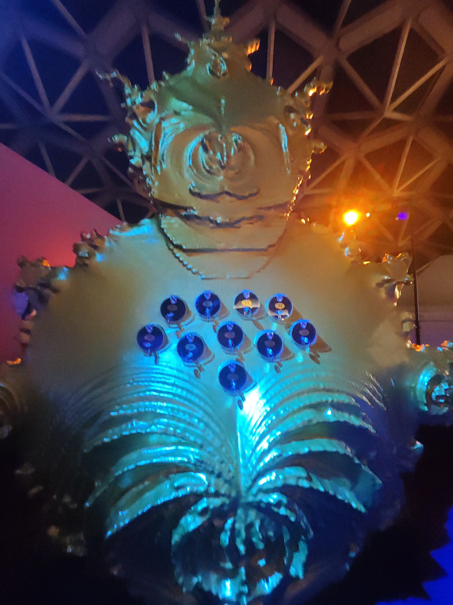 The image shows a large, ornate, golden sculpture that resembles a seashell or a crown with a face-like structure at the top. The sculpture is illuminated with a mix of blue and orange lights, creating a theatrical and somewhat eerie atmosphere. Several small, circular objects, possibly lights or decorative elements, are embedded in the chest area of the sculpture. The background appears to be an indoor space with a geodesic dome-like ceiling structure and pink and orange lighting. The sculpture is located at the entrance of the Evoluon in Eindhoven, Netherlands. The Evoluon is a science museum housed in a distinctive UFO-shaped building, which was originally built as a Philips pavilion for the 1964 Brussels World's Fair.