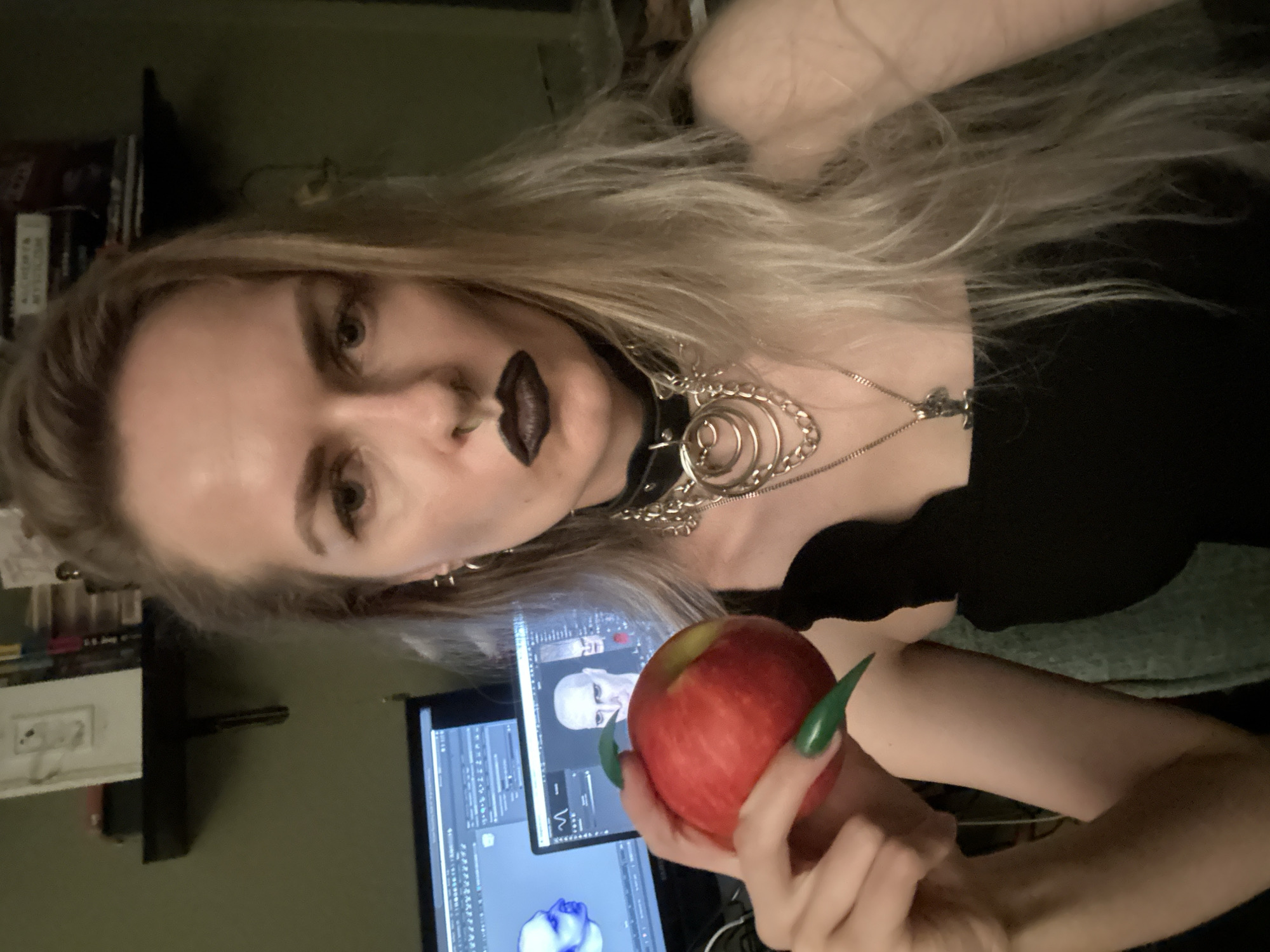 A fair-skinned person with long blonde hair and black lipstick holds a red apple. The person wears a black top, a choker with metal rings, and a necklace with a pendant. The person has pierced ears and a nose ring. The nails on the person's hand holding the apple are long and green. In the background is a computer monitor displaying a 3D model of a human head. Shelves with books are also visible in the background. There is no information in the image to determine a location in the city of Cape Coral, United States of America.