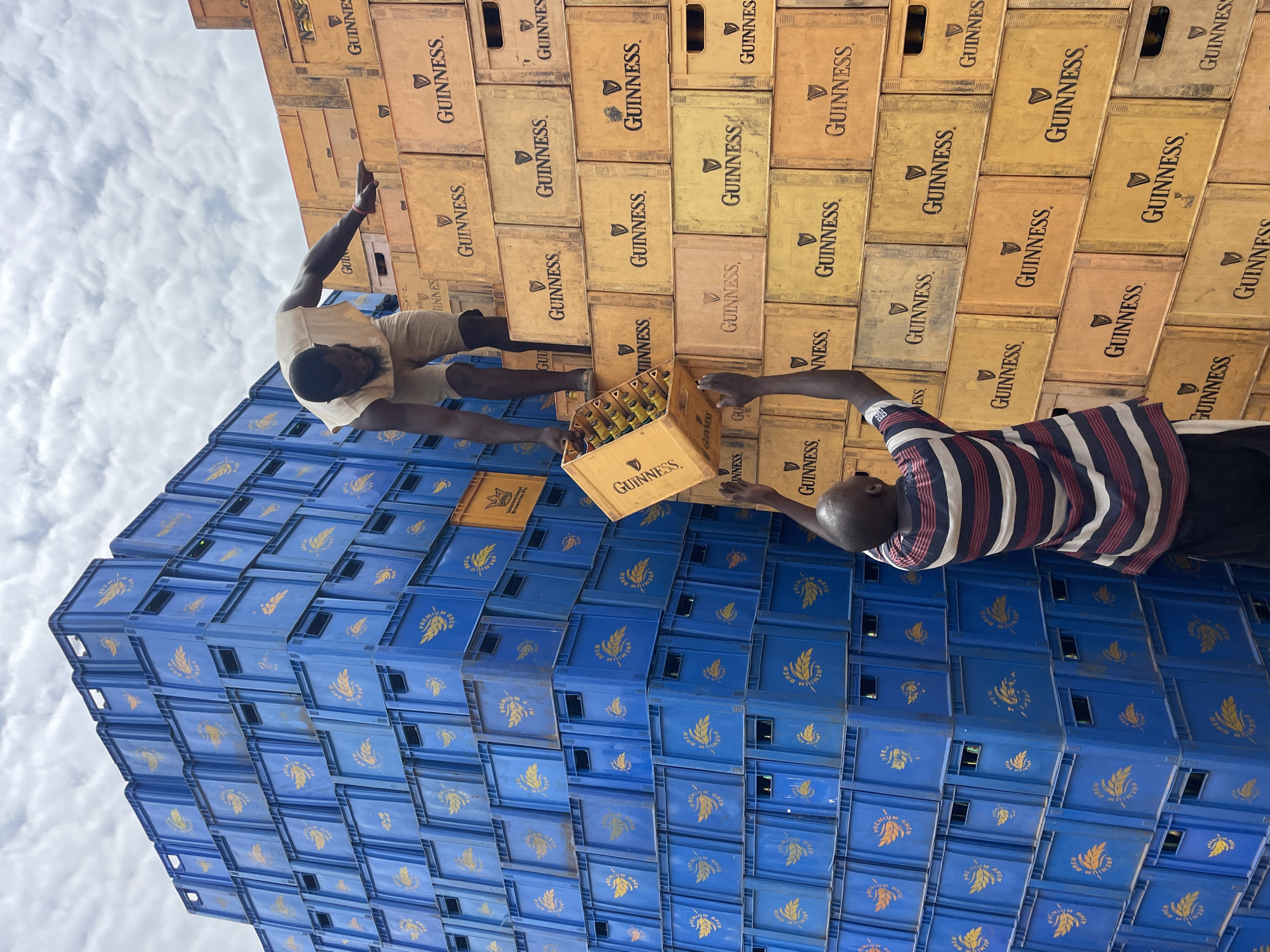 The image shows two men near a stack of beer crates in Bukuru, Nigeria. The stack is divided into two sections: the lower section is composed of blue crates, while the upper section is made of brown crates labeled "Guinness". One man, wearing a light-colored outfit, is standing atop the crates with his arm extended upwards. The other man, wearing a striped shirt, is handing him a crate of Guinness beer. The sky is visible in the background.