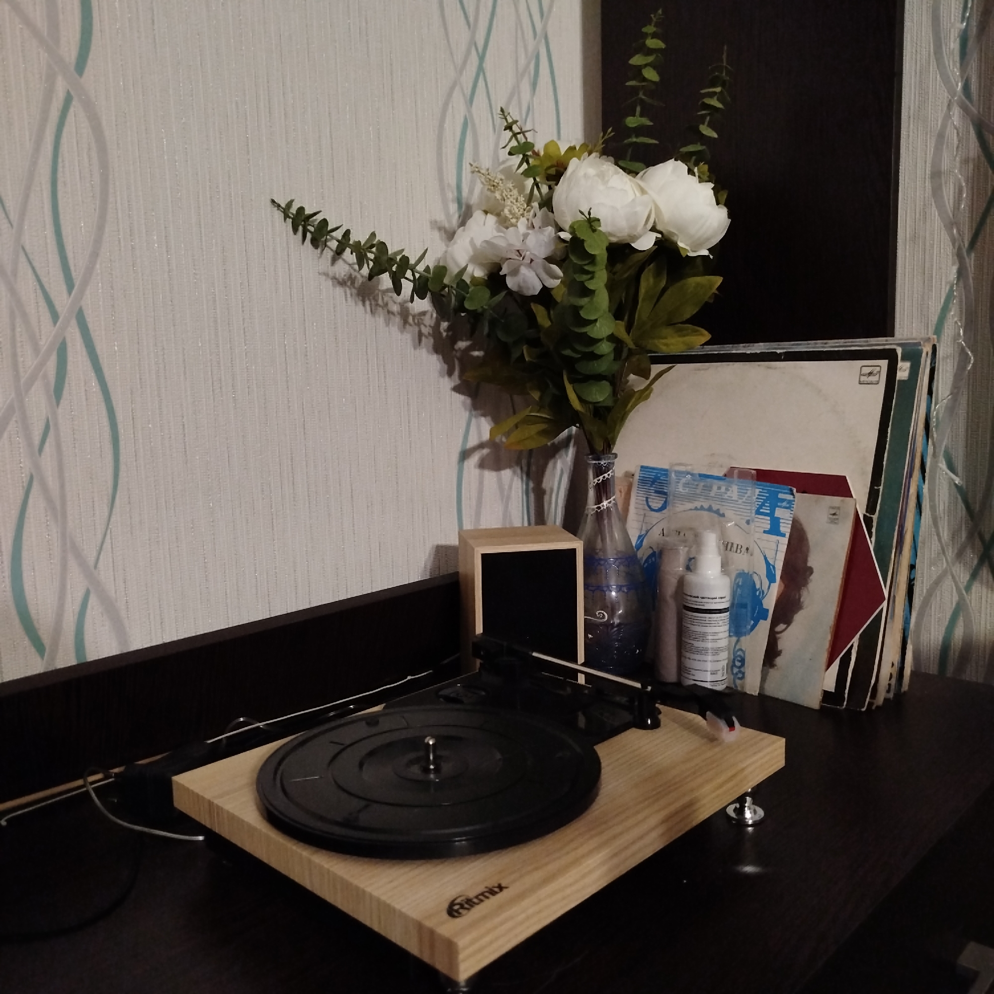 The image depicts an indoor setting featuring a Ritmix record player placed on a dark-colored surface, likely a desk or shelf. Positioned behind the record player are a vase filled with white flowers and greenery, a small wooden box with a black interior, and a stack of vinyl records in their sleeves. A bottle of white liquid with black labels is also present. The background consists of a wall with a pattern of white and light blue waves, along with a dark rectangular shape, potentially a piece of furniture or art.