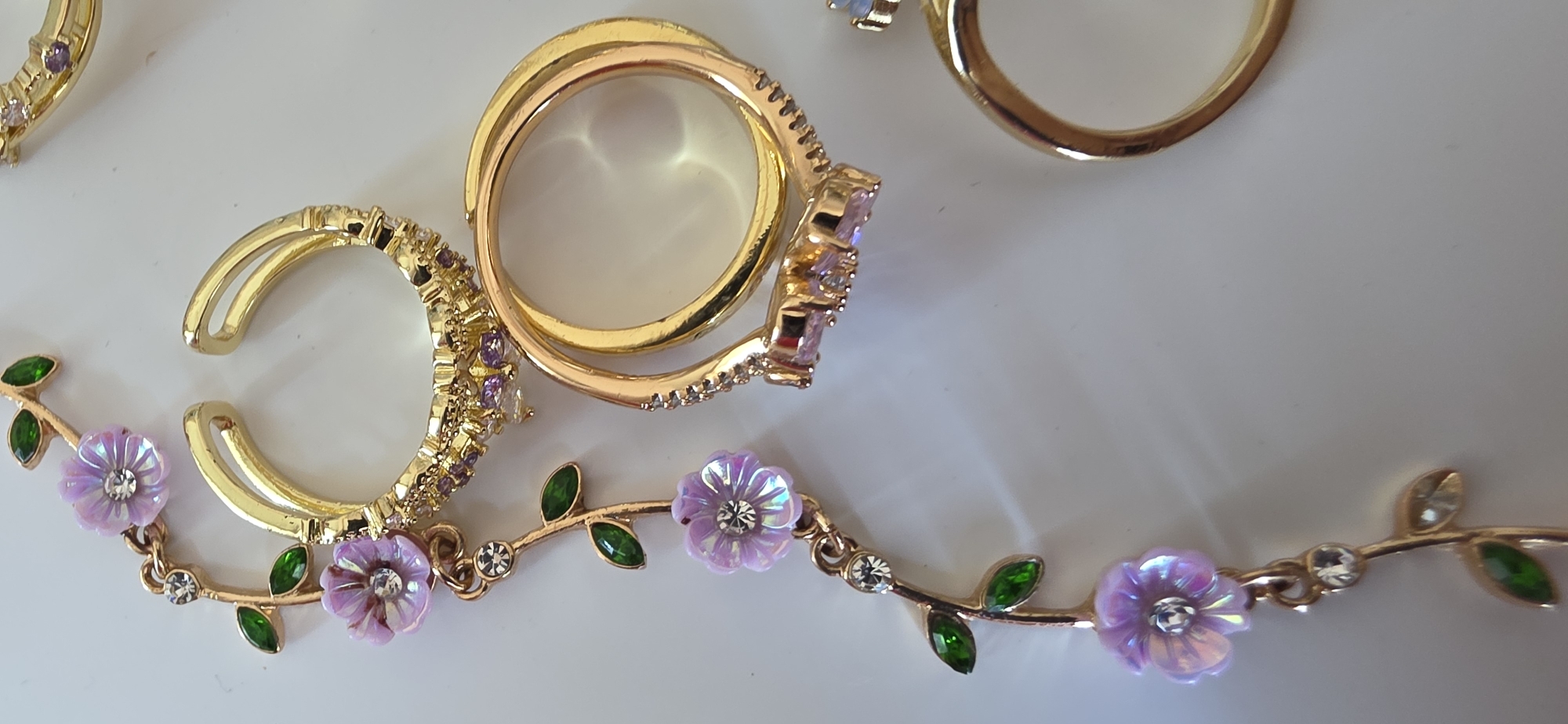A close-up, overhead shot displays a collection of gold-colored jewelry pieces, featuring floral and leafy motifs, arranged on a white surface. The jewelry includes a dangling chain with small green enamel leaves and iridescent purple flowers, each adorned with a clear crystal at its center. Several rings are also present. One ring is a double-banded design with clear and purple cubic zirconia. Another ring is a simple gold band, and a third, partially visible, appears to be another gold band. The lighting is bright, creating reflections on the polished metal and stones. The background is a plain white surface, likely a table or countertop. There are no people visible in the image, and no indication of the specific location within Sumgayit, Azerbaijan.