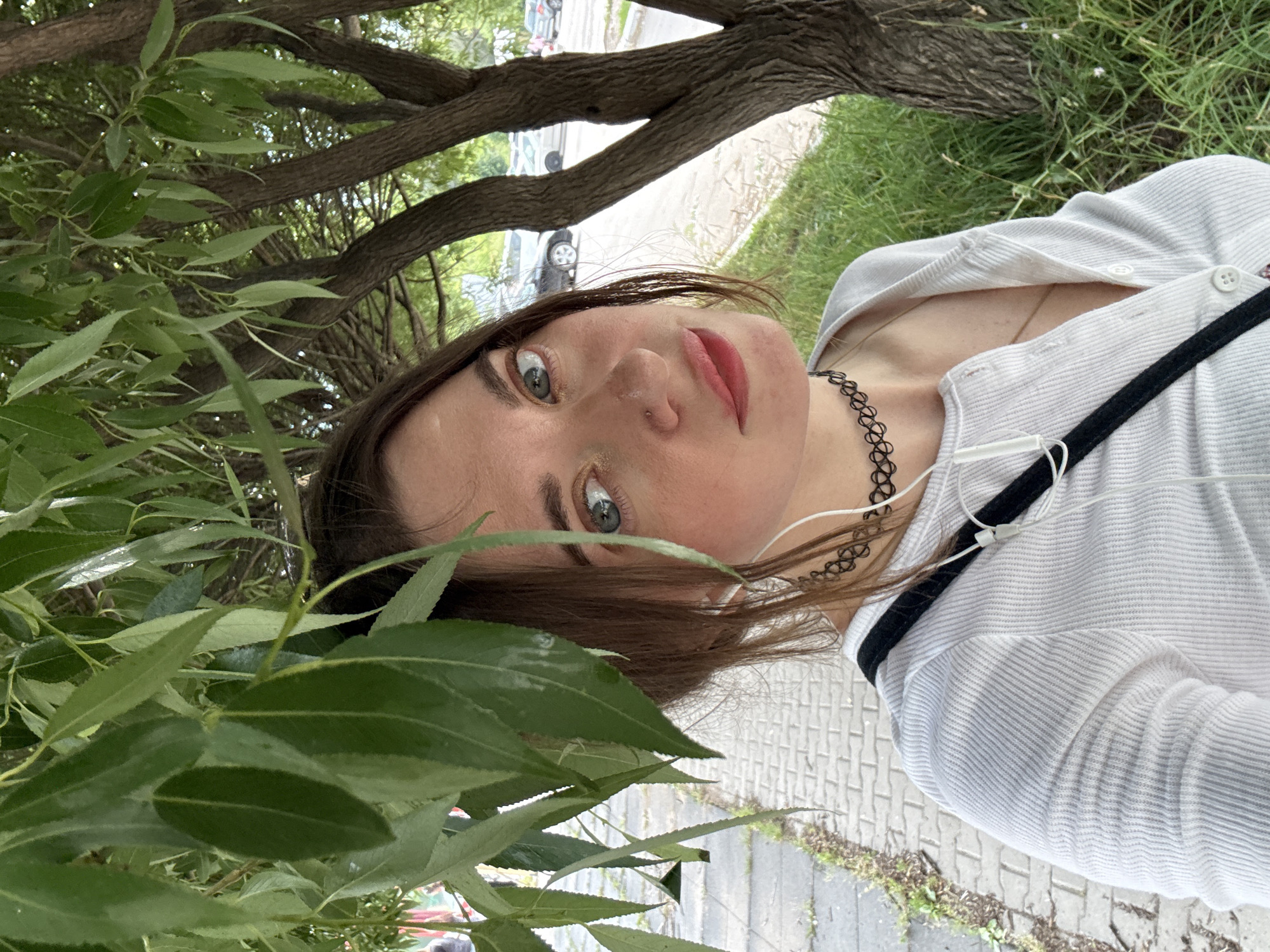 A young woman with light brown hair and green eyes takes a selfie outdoors under the shade of a tree. She is wearing a white, long-sleeved, ribbed shirt with a low neckline and a black choker necklace. White earphones are visible, plugged into her shirt. The background shows a sidewalk, grass, tree trunks, and parked cars on a street. The lighting suggests it is daytime.