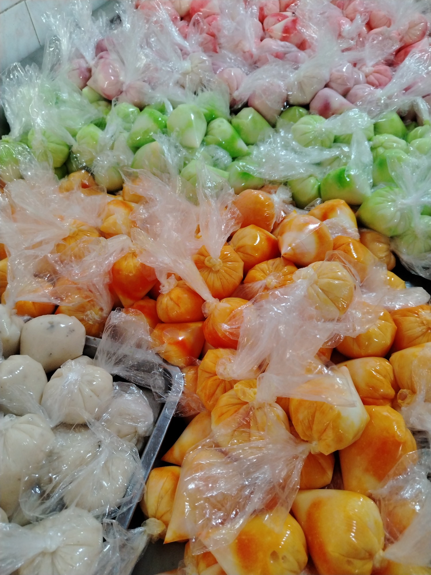 The image depicts a collection of wrapped sweets or desserts in a commercial or retail setting, likely in Tinaquillo, Venezuela. These appear to be individual portions contained in clear plastic bags. The items are assorted in color: pink, green, orange, and white. There are no people visible in the image. The overall environment suggests a point of sale or storage area.