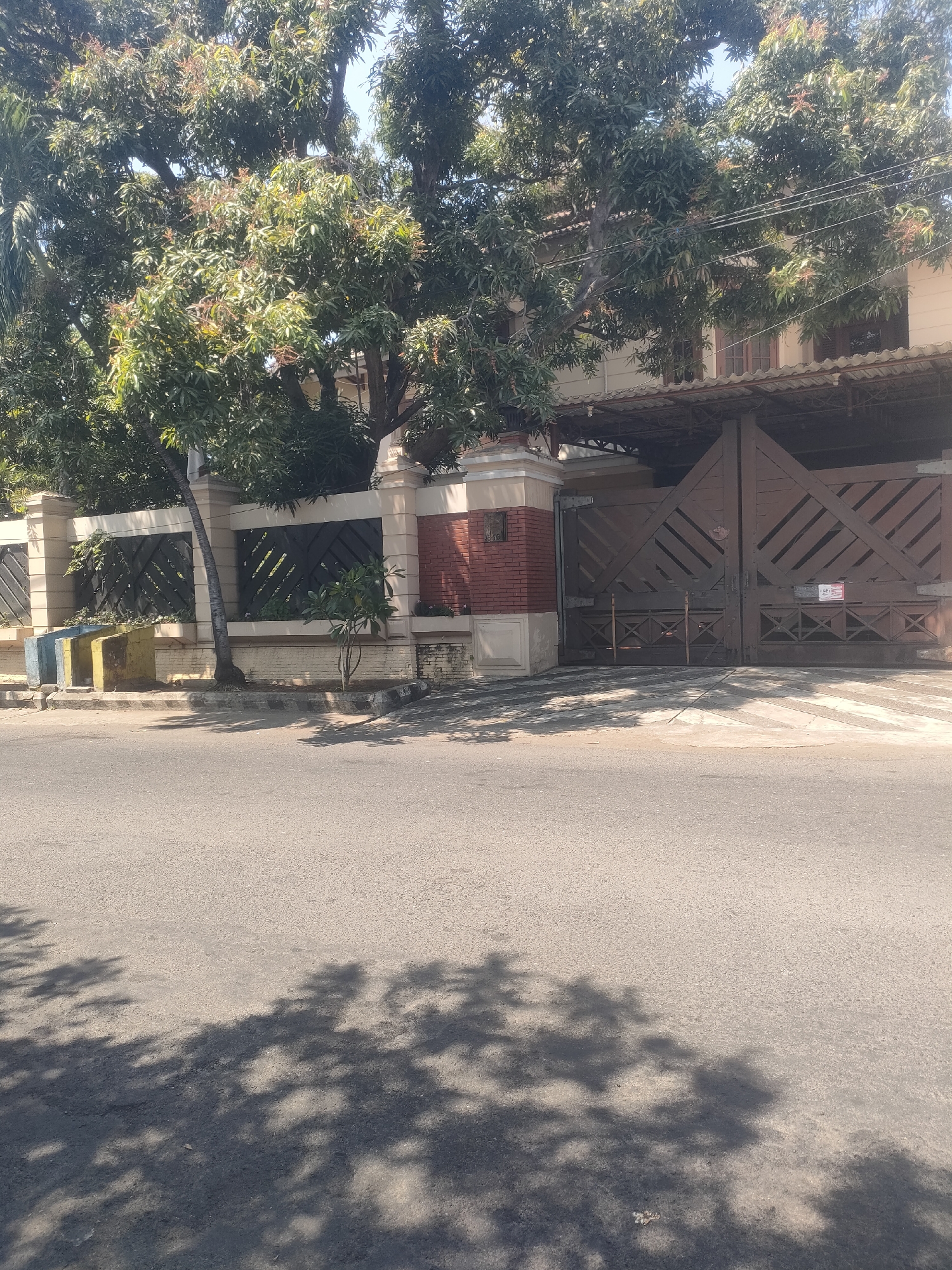 The image shows a wide, paved street in front of a residential property in Surabaya, Indonesia. The property is enclosed by a decorative fence and a large, wooden gate. A mature tree with dense foliage stands in front of the fence, and its branches extend over the gate and the house. The house itself appears to be a multi-story structure with a light-colored facade and brown-trimmed windows. The gate is made of dark wood with a geometric pattern and has a small sign on it. The street is empty of vehicles and people, and the sunlight casts shadows of the tree's leaves onto the asphalt. The scene suggests a quiet, sunny day in a residential area of Surabaya.