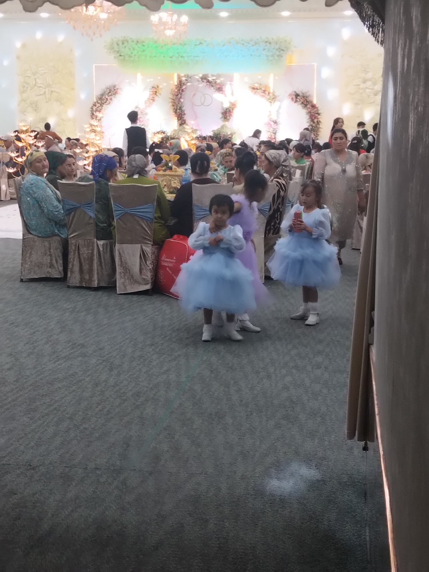 The image depicts a formal indoor gathering, likely a wedding reception, held in the city of Siyozi-poyon, Uzbekistan. A large room is set up with numerous chairs draped in brown covers and adorned with blue sashes. In the background, a decorated archway with floral arrangements and illuminated elements suggests a ceremonial area. Several adults are present, seated and standing, some wearing traditional headscarves. In the foreground, two young girls in voluminous light blue dresses stand on a grey carpeted floor, one holding a red object. The overall ambiance suggests a celebratory event.