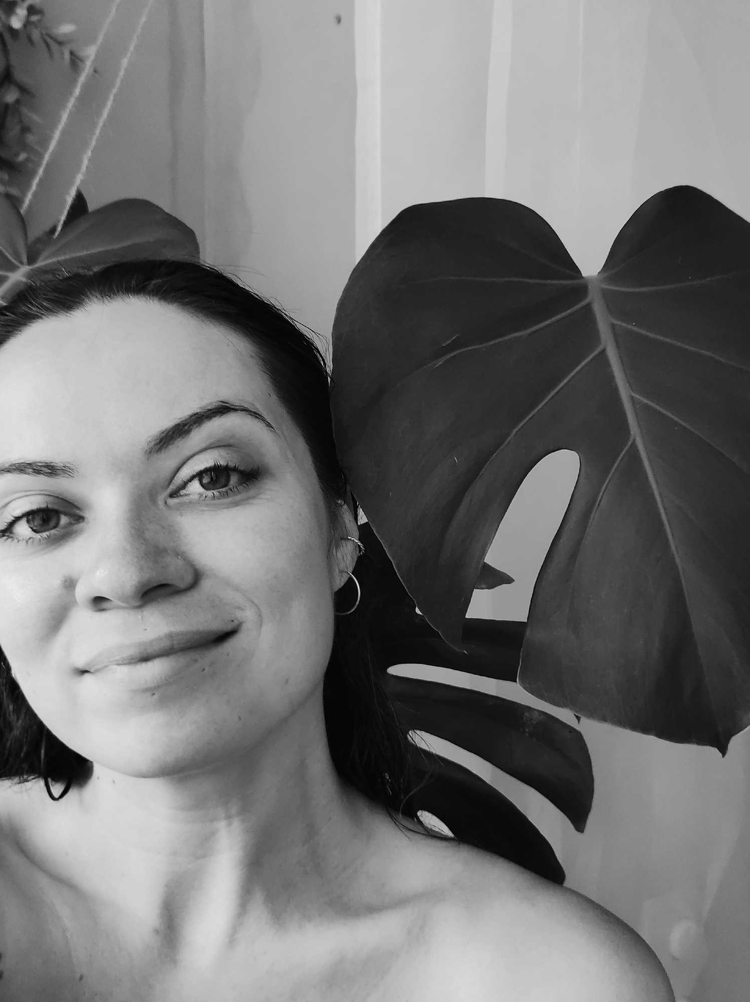 A monochrome close-up photo features a woman and a large leaf. The woman, likely in her late 20s or early 30s, is positioned on the left, facing the camera with a slight smile. Her hair is dark and appears wet. She is wearing hoop earrings. Her shoulders are bare.
To her right is a large, prominent Monstera leaf with a characteristic split. The background is a plain white or light-colored wall.
The photo's location in the city of Chaikovsky, Russia, cannot be determined from the visual information provided.