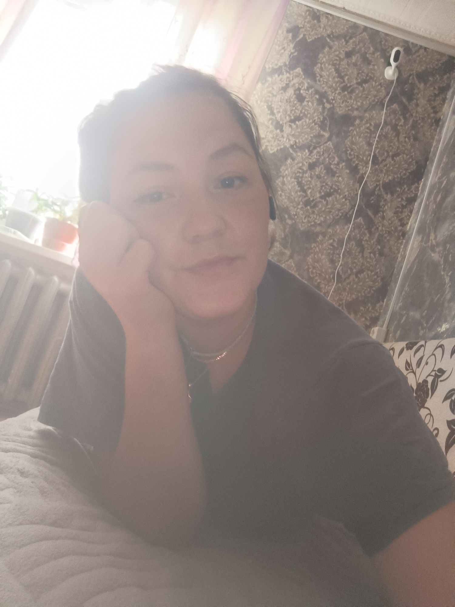 The image is a selfie taken by a person with dark hair, resting their head on their hand. They are wearing a dark grey t-shirt and a silver necklace. In the background, there is a window with light coming through, some potted plants, a radiator, patterned wallpaper, and a person in a couch with a floral pattern. There are also white earphones with a wire visible. The scene appears to be indoors, likely a home environment. The image quality is not very high, and the lighting is strong, creating a glare. There is no visible indication of the specific location within the city of Karsha, Russia.