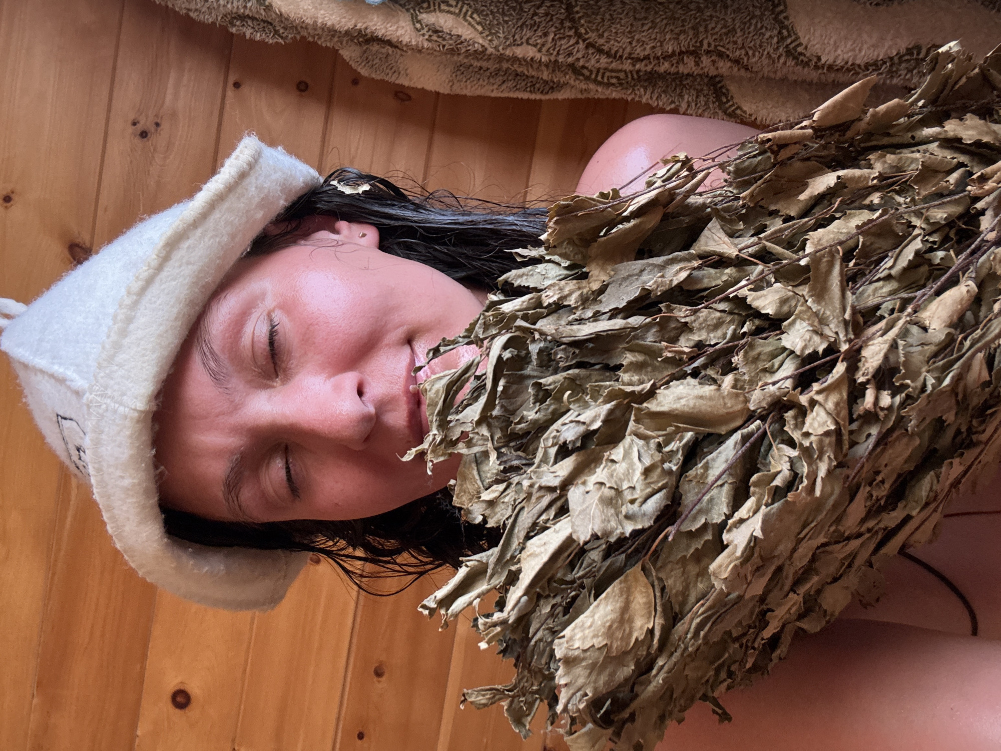 A person is reclining on wooden planks, possibly in a sauna, with a white cap on their head. They have dark, wet hair and their eyes are closed. A bundle of dried leaves and branches, resembling a venik (sauna whisk), is near their face and upper body. The background suggests the interior of a sauna, with wooden walls and a towel visible above. The location might be Chaikovsky, Russia.