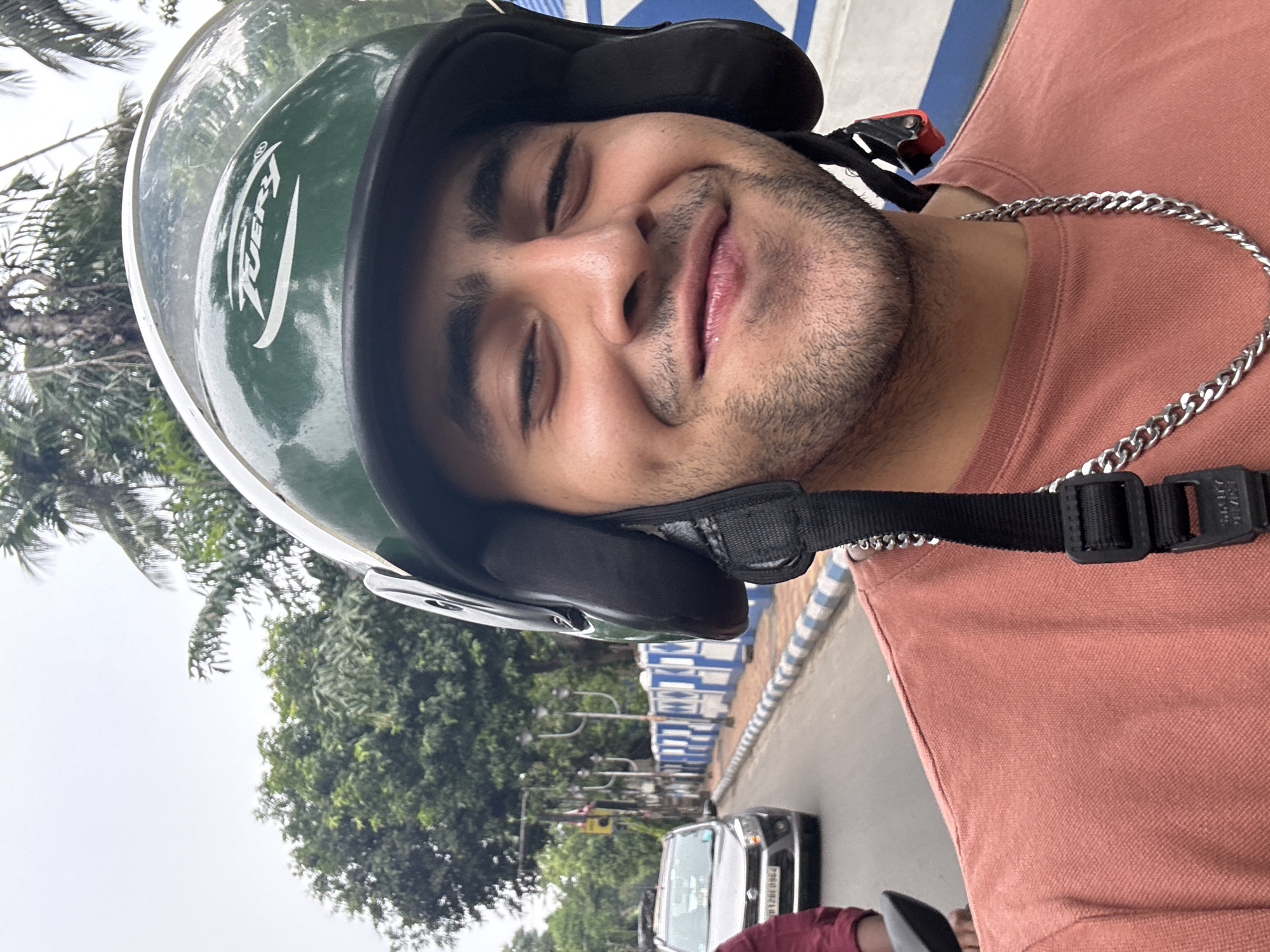 This is a close-up portrait of a young man, likely taken as a selfie or by someone riding alongside. He is wearing a dark green helmet with a clear visor, on which the brand name "TVERY" is clearly visible. The man has a light beard or stubble and is smiling warmly with his eyes partially closed, conveying a happy and relaxed expression. He is wearing a pinkish-orange t-shirt and a silver curb chain around his neck. Part of the helmet's chin strap buckle is visible, showing the partially legible text "SMIT/DRIVER".
The setting is an outdoor street scene in Kolkata, India, during the daytime, under what appears to be a bright, possibly overcast, sky. In the background, lush green trees, including palm trees, are visible. A road runs alongside, featuring a black hatchback car with a partially visible license plate ("P12BCQ8847" can be deciphered). A blue and white striped barrier or divider lines the edge of the road, a common sight in urban Indian environments. The presence of the helmet strongly suggests the man is riding a two-wheeler.